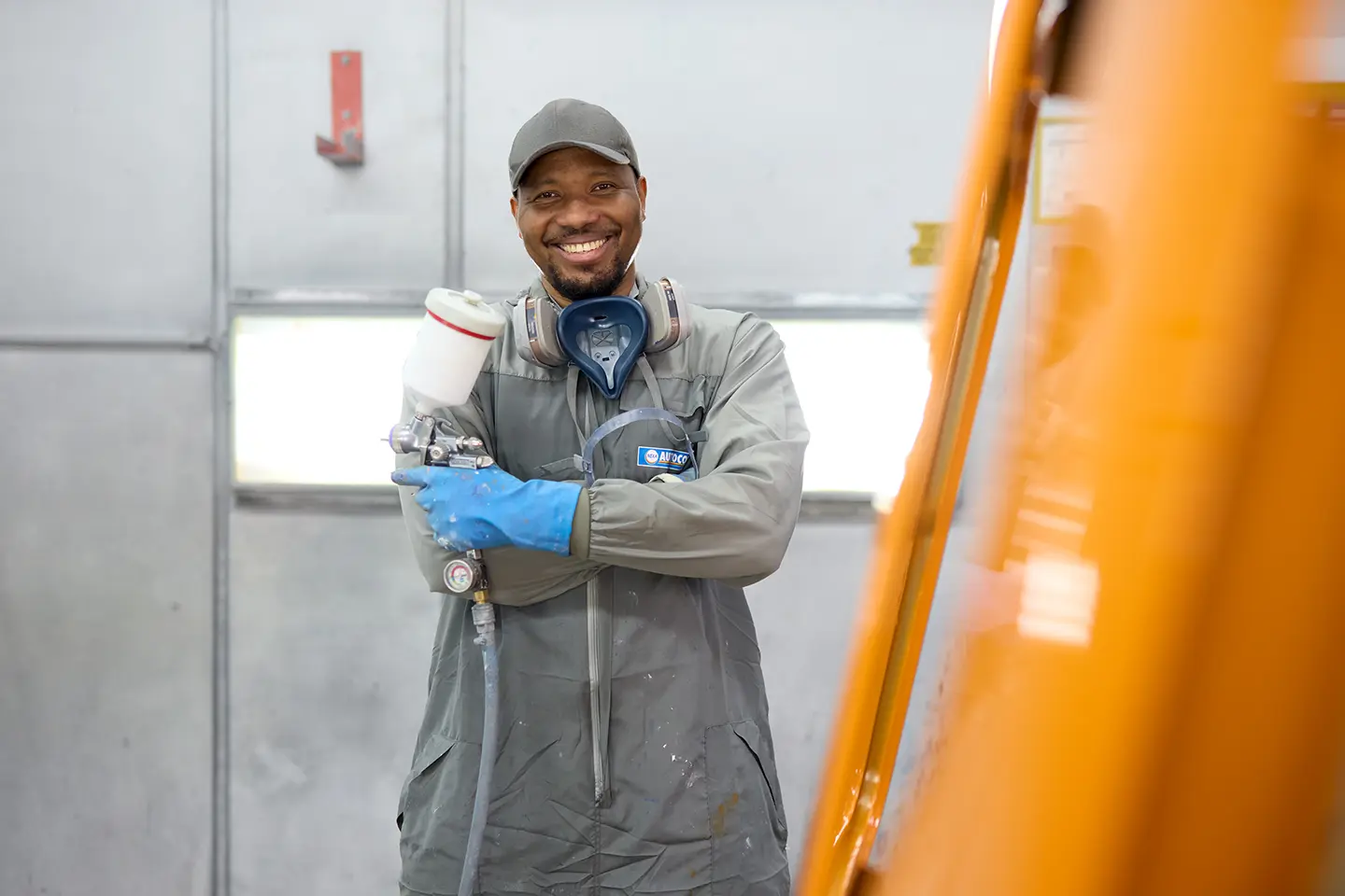 Lächelnder stolzer Mitarbeiter in grauer Schutzkleidung, blauen Handschuhen und Kappe hält eine Lackierspritzpistole in einer Werkstatt.