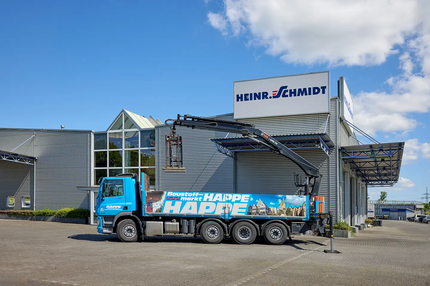 Blauer Baustoff-LKW mit Kran vor einem Gebäude mit HEINR. SCHMIDT-Schild bei klarem Himmel.