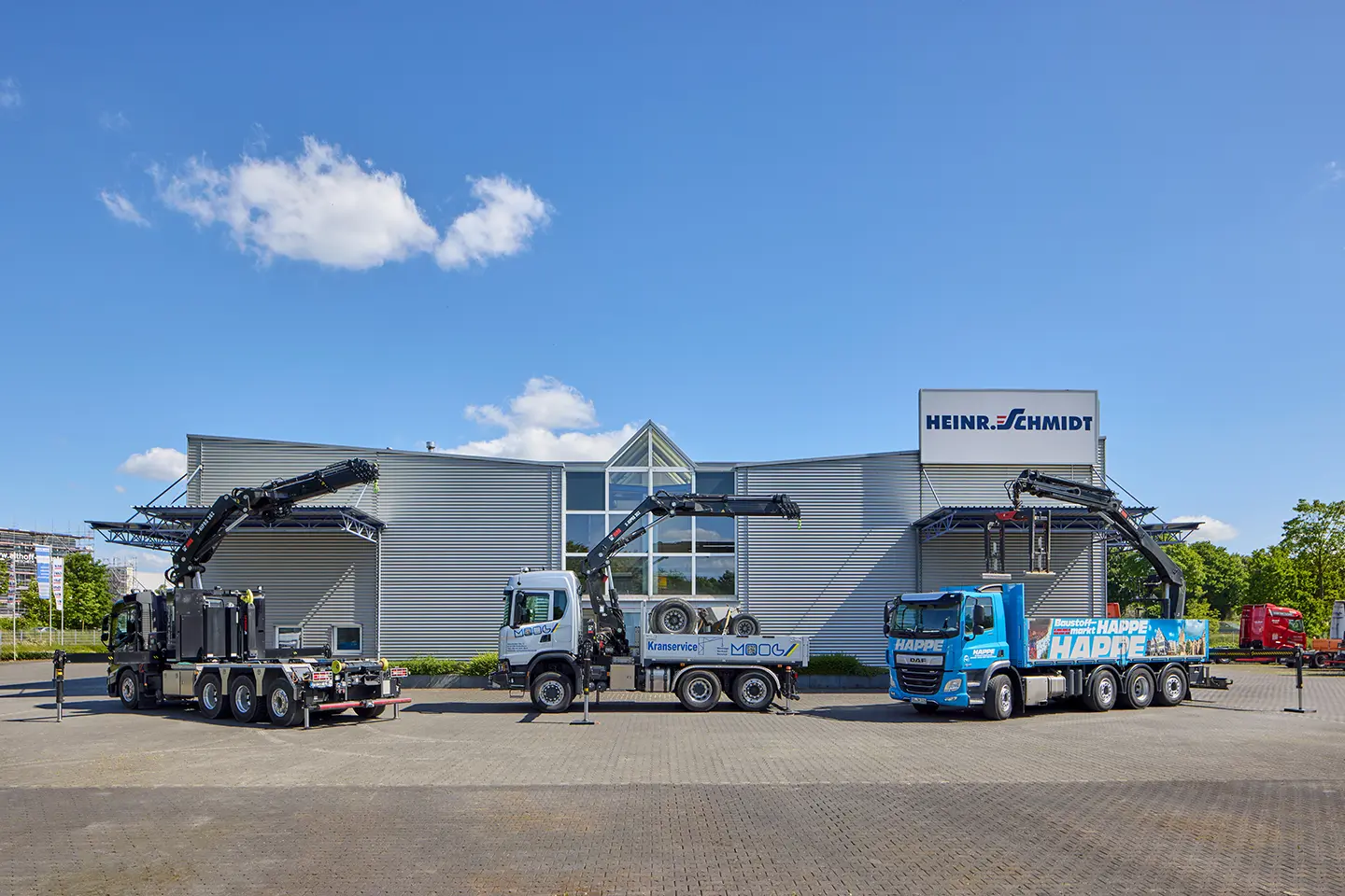 Drei Lkw mit Hebekranen stehen vor einem modernen Gebäude mit dem Schild 'HEINR. SCHMIDT' unter blauem Himmel.