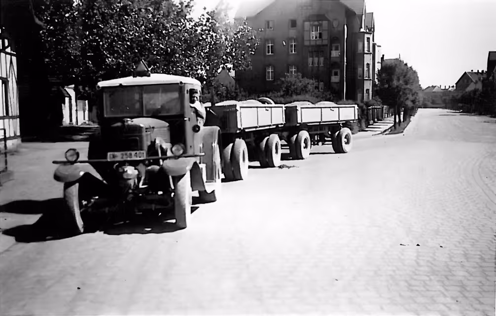 Schwarz-weiß-Foto eines historischen Lastwagens mit zwei Anhängern auf einer gepflasterten Straße in einer Wohngegend.