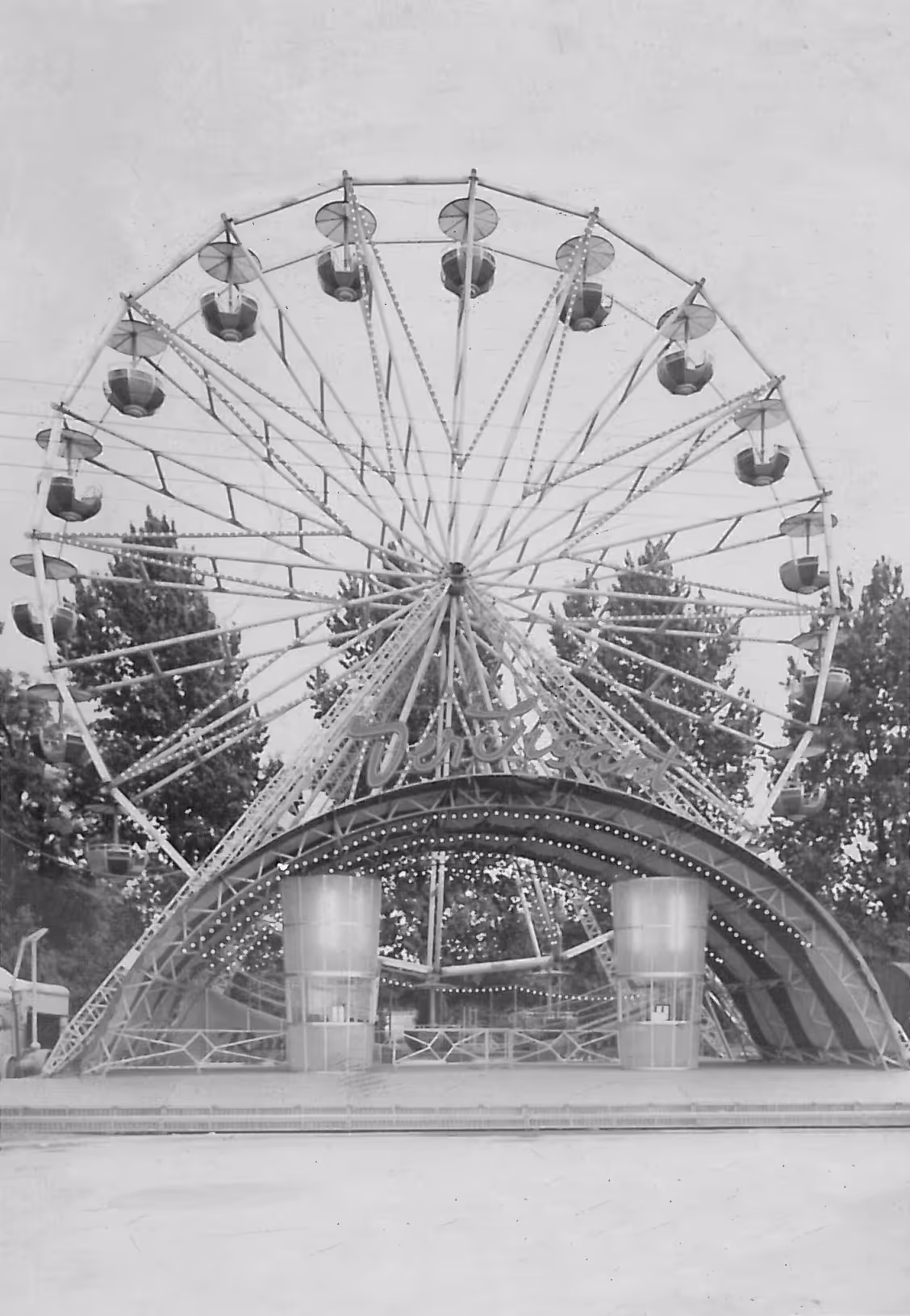 Schwarz-Weiß-Foto eines großen Riesenrads mit geschlossenen Gondeln und einem beleuchteten Eingangsbereich, dahinter Bäume.