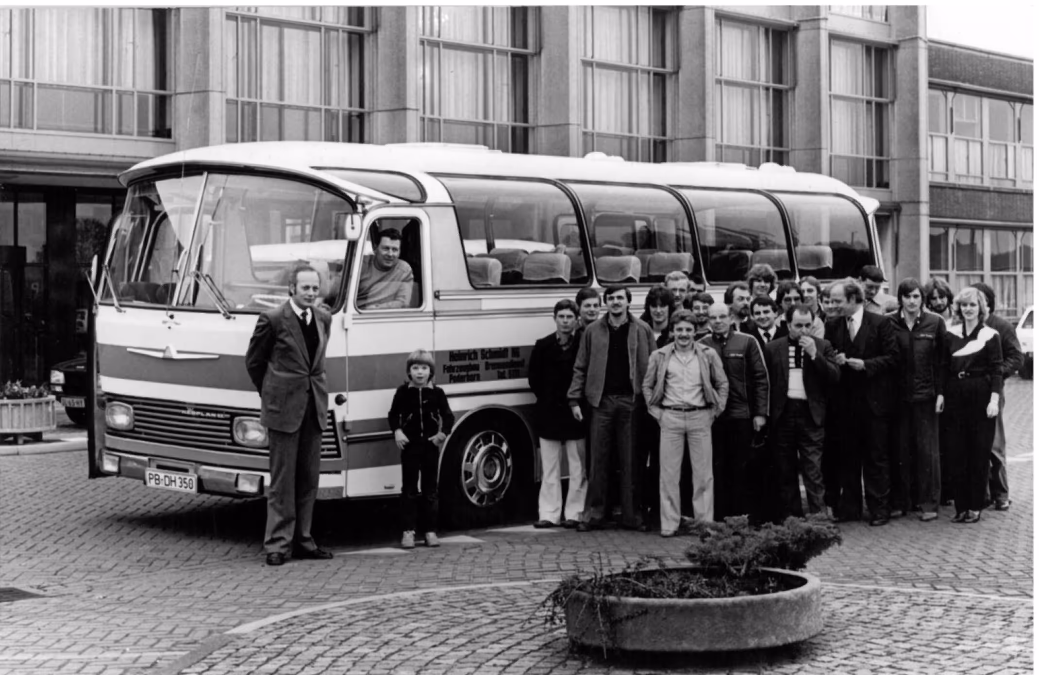 Schwarz-weiß-Foto einer großen Gruppe von Menschen, die vor einem alten Reisebus auf einem gepflasterten Platz stehen.