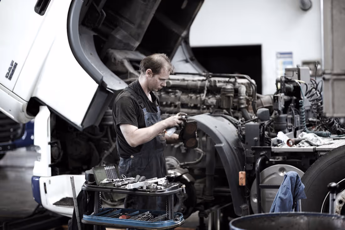 Mechaniker überprüft ein Teil vor einem geöffneten Lkw-Motor, mit Werkzeugwagen im Vordergrund.
