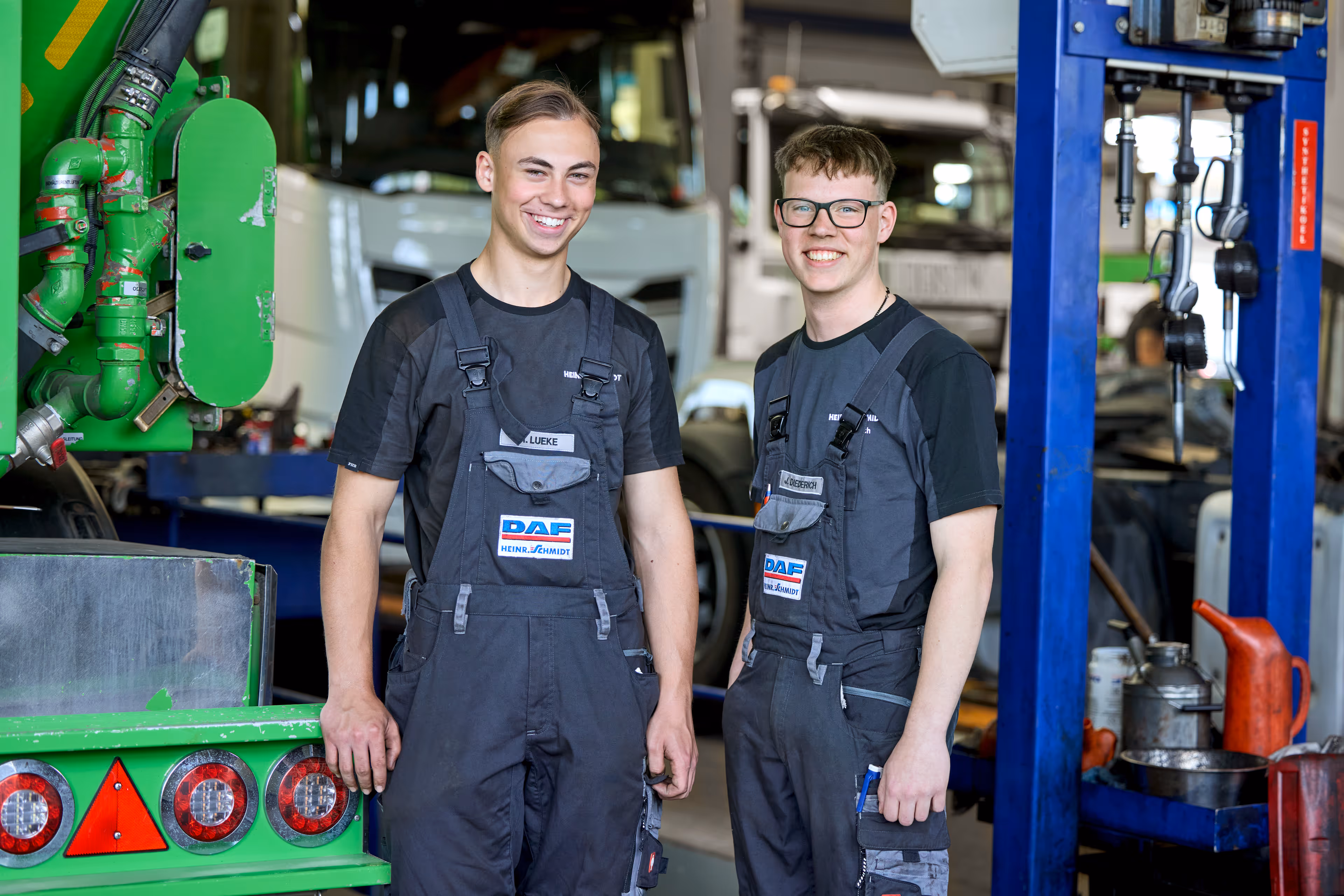Zwei junge Männer in Arbeitskleidung stehen lächelnd in einer Werkstatt mit Fahrzeugen und Werkstattgeräten im Hintergrund.