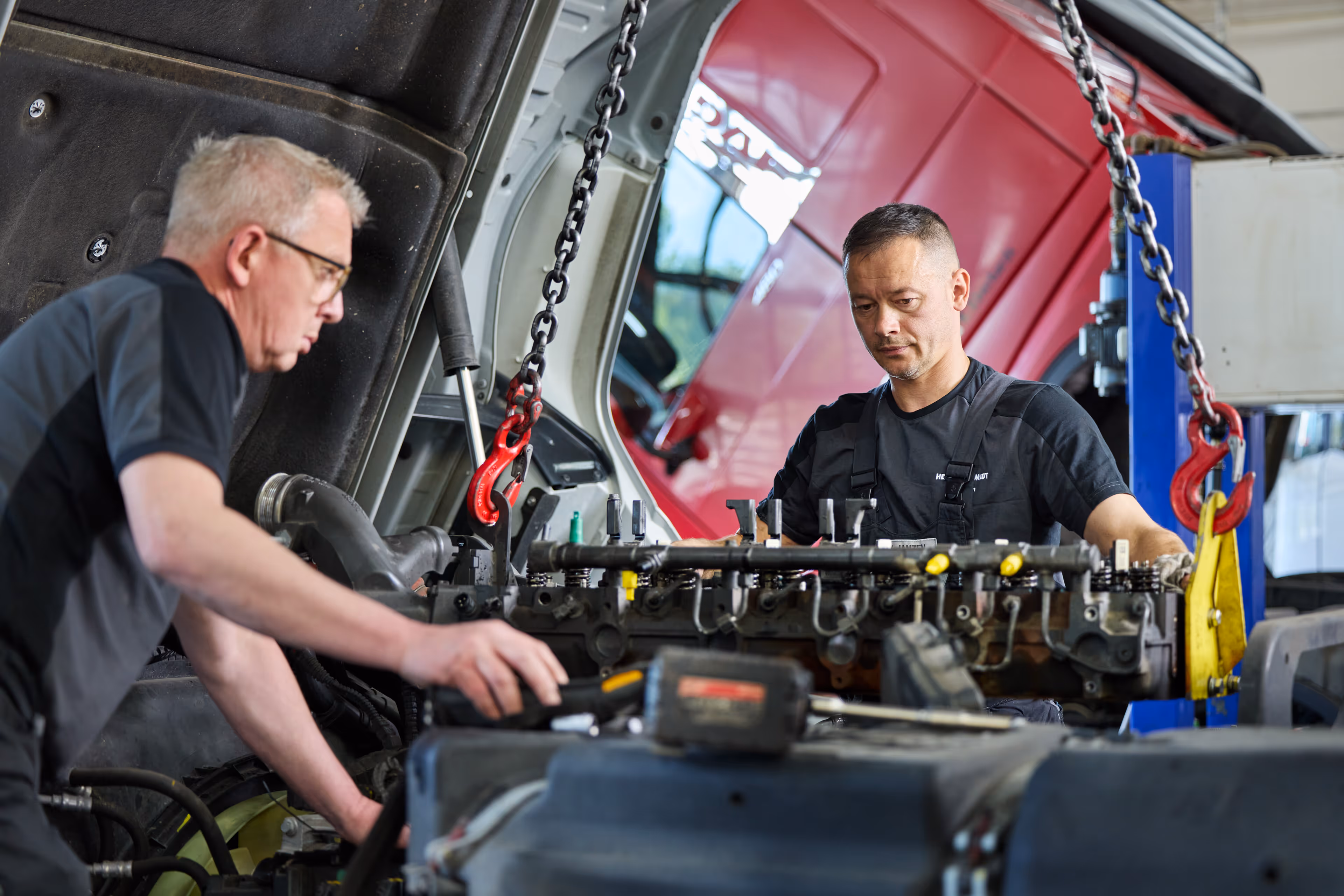 Zwei Mechaniker arbeiten gemeinsam in einer Werkstatt an einem Motor, der mit Ketten an einem Hebezeug befestigt ist.