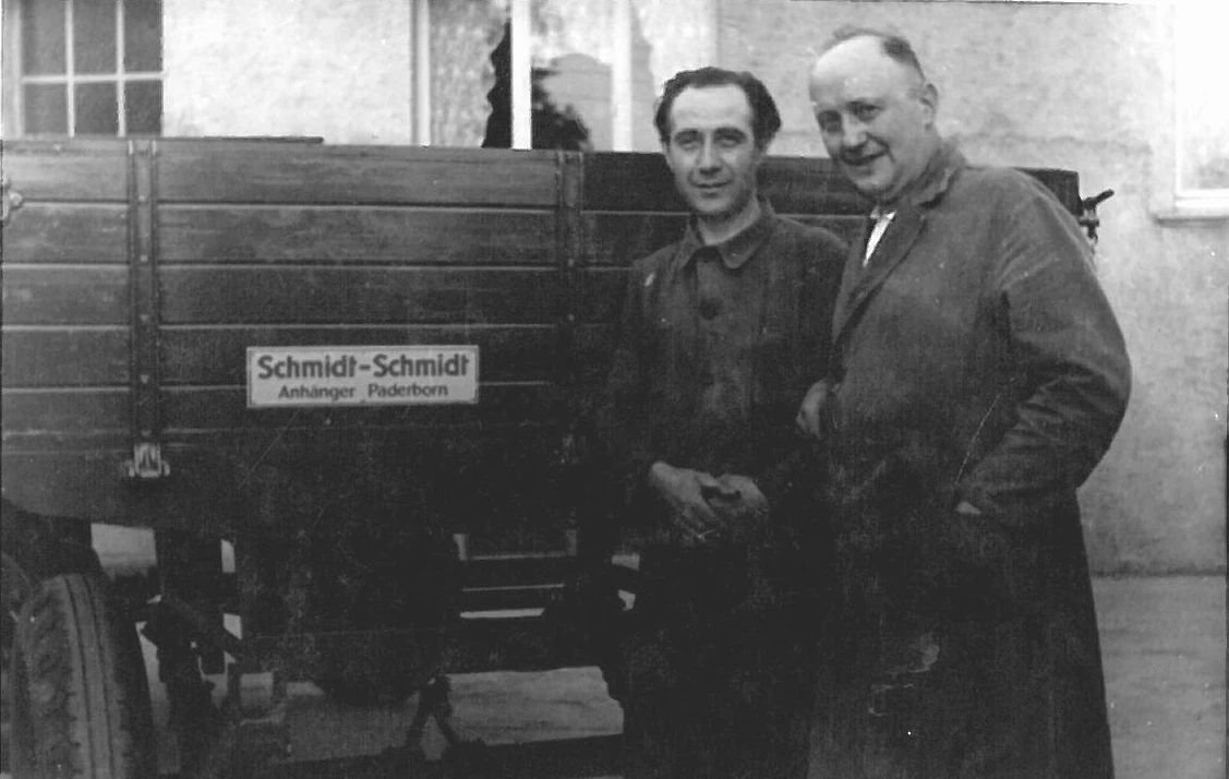 Zwei Männer in Arbeitskleidung stehen vor einem Anhänger mit dem Schild 'Schmidt–Schmidt Anhänger Paderborn'.