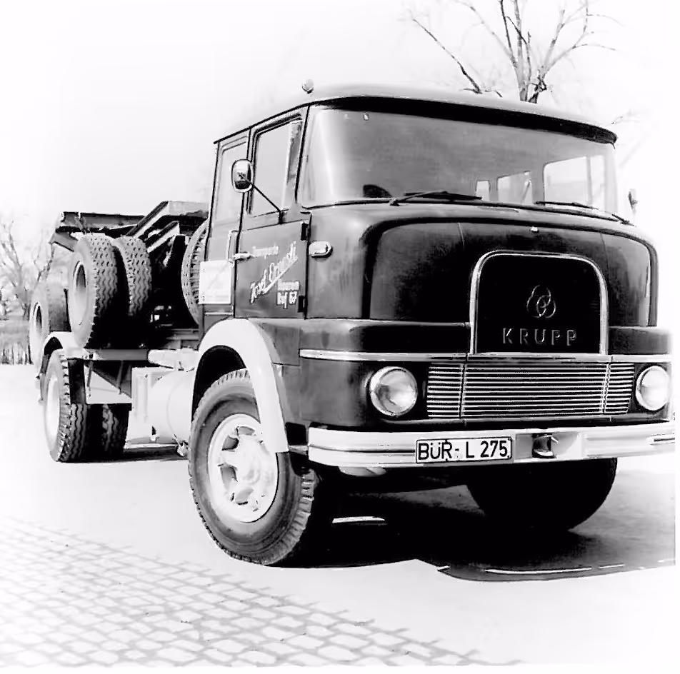 Schwarz-weiß Foto eines historischen Krupp Lkw mit zwei Ersatzreifen auf einem Kopfsteinpflaster.
