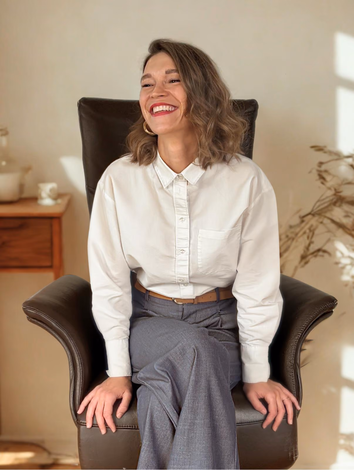 Femme souriante assise sur un fauteuil en cuir marron avec chemise blanche et pantalon gris.