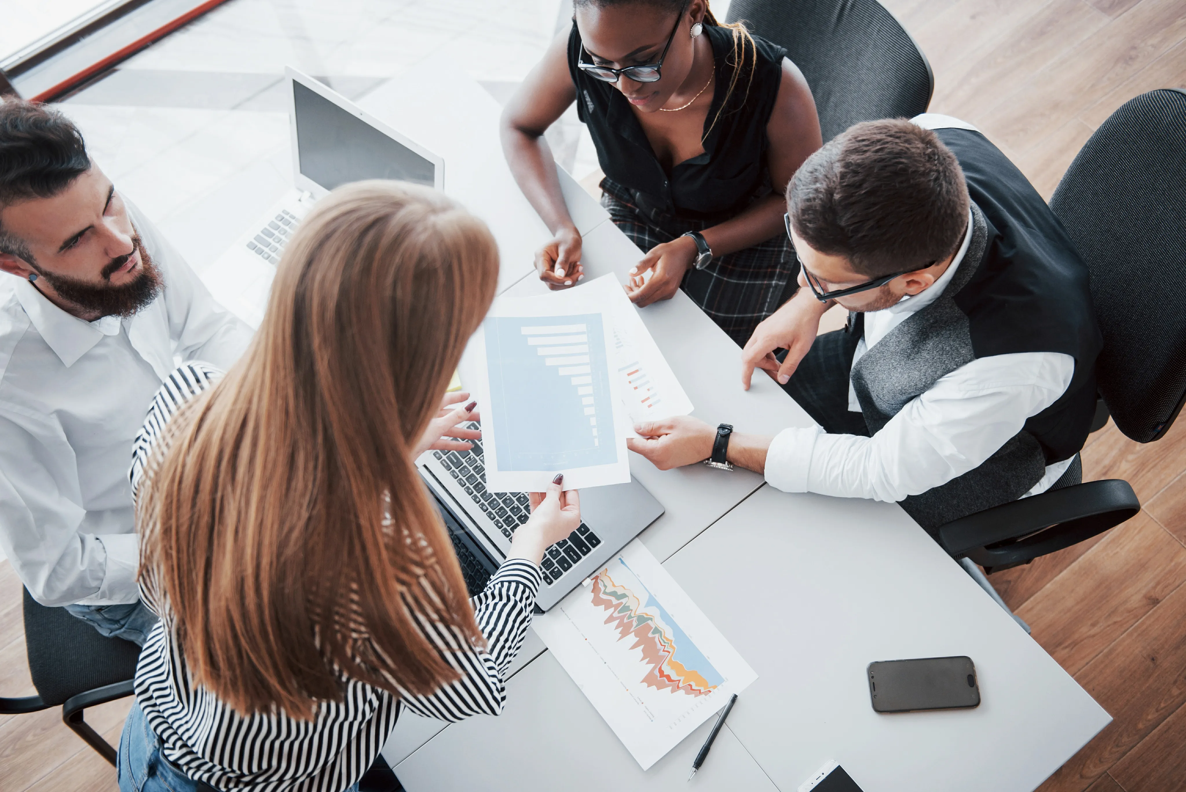 Four coworkers gathered around a table, reviewing charts and graphs together, with laptops and documents in view.
