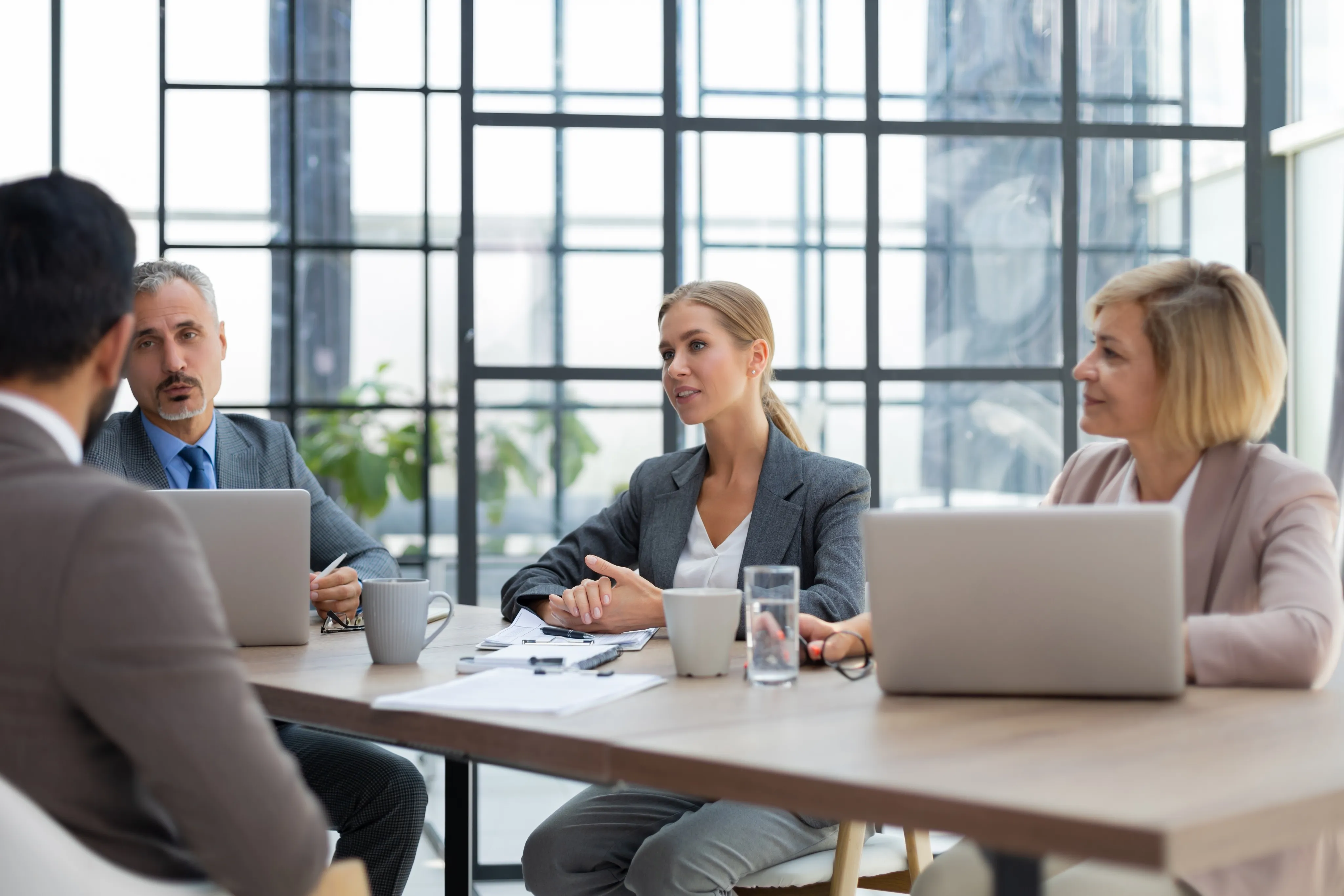 Three business professionals in a modern office having a meeting, sitting at a table with laptops and coffee mugs, facing another person.