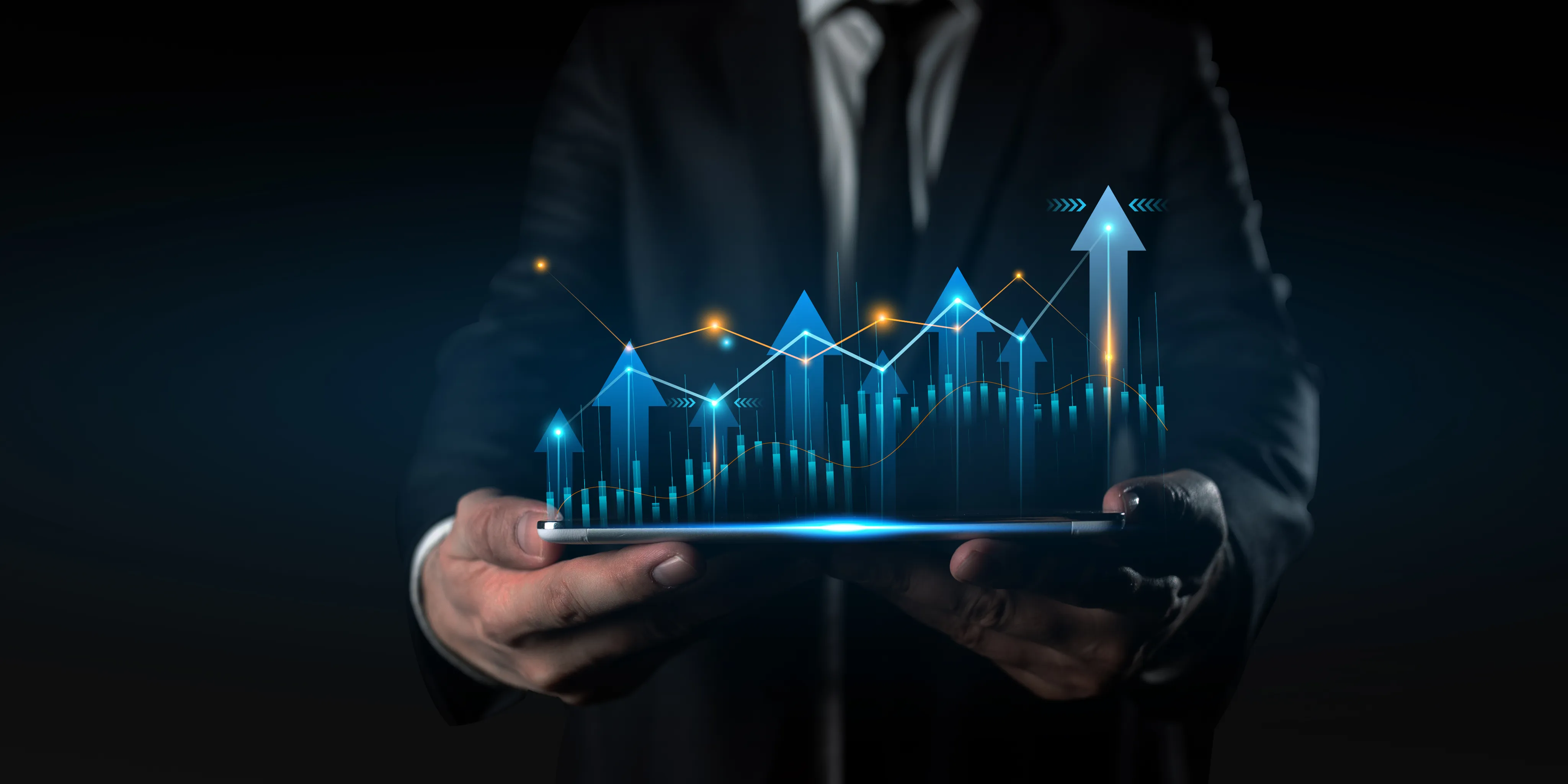 A person in a suit holding a tablet with digital blue arrows and graphs rising upward, representing financial or business growth.