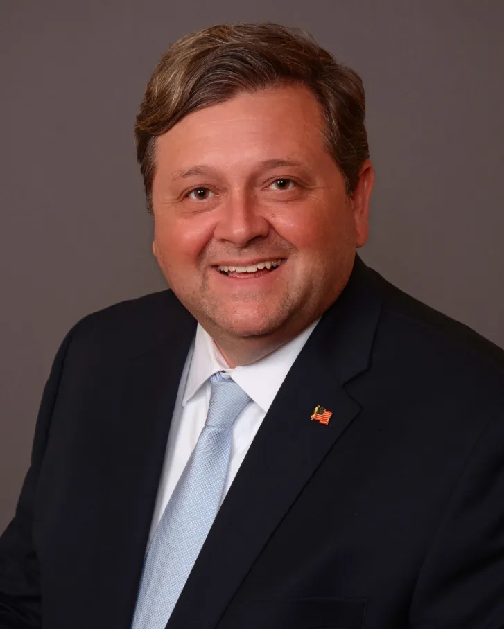 A man in a suit and tie smiling, wearing a small U.S. flag pin on his lapel, in front of a neutral gray background.