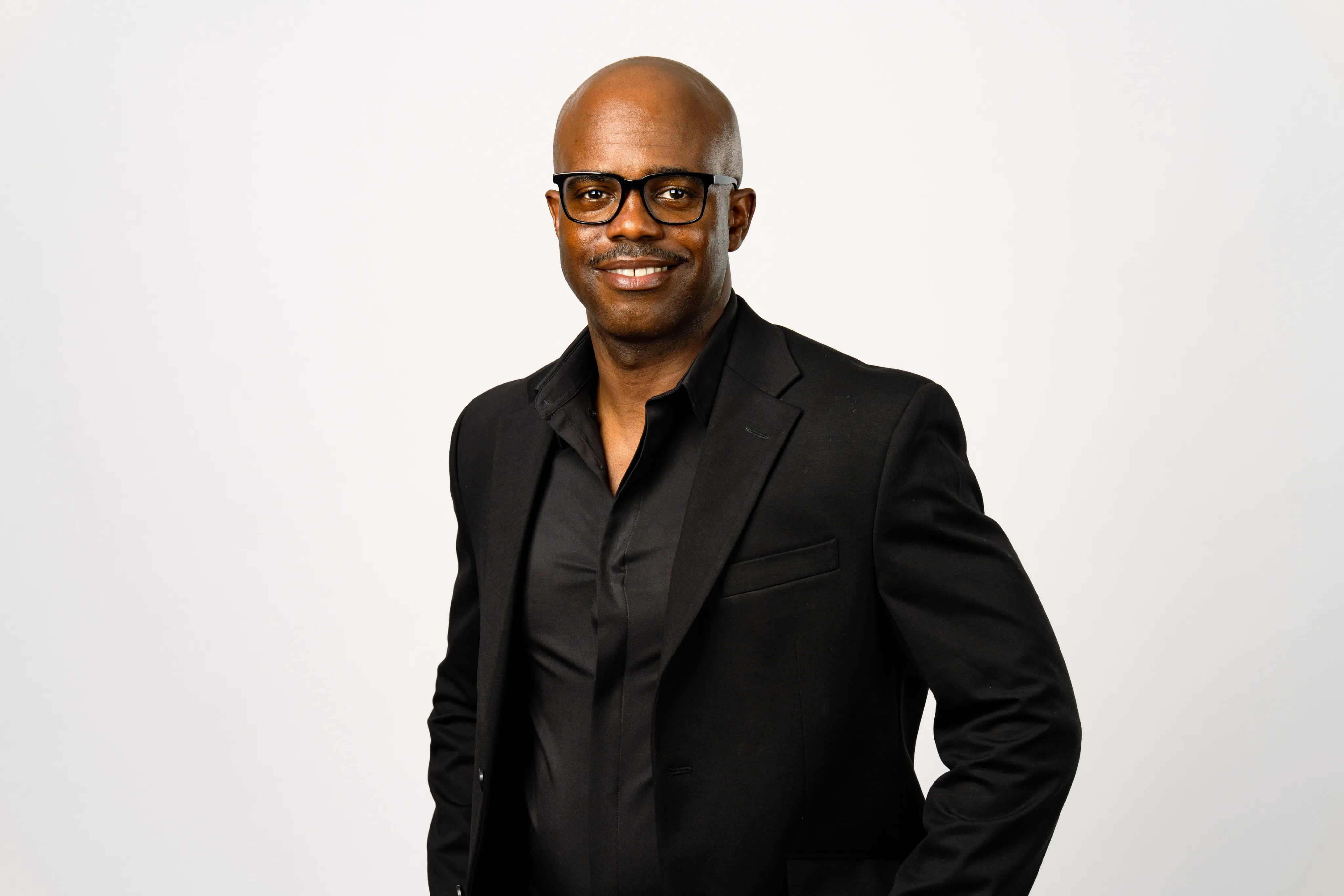 A man wearing glasses and a black suit jacket, smiling, standing against a plain light background.