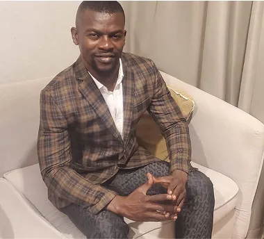 A man in a plaid suit jacket and patterned pants sitting on a light-colored couch, smiling with hands clasped.