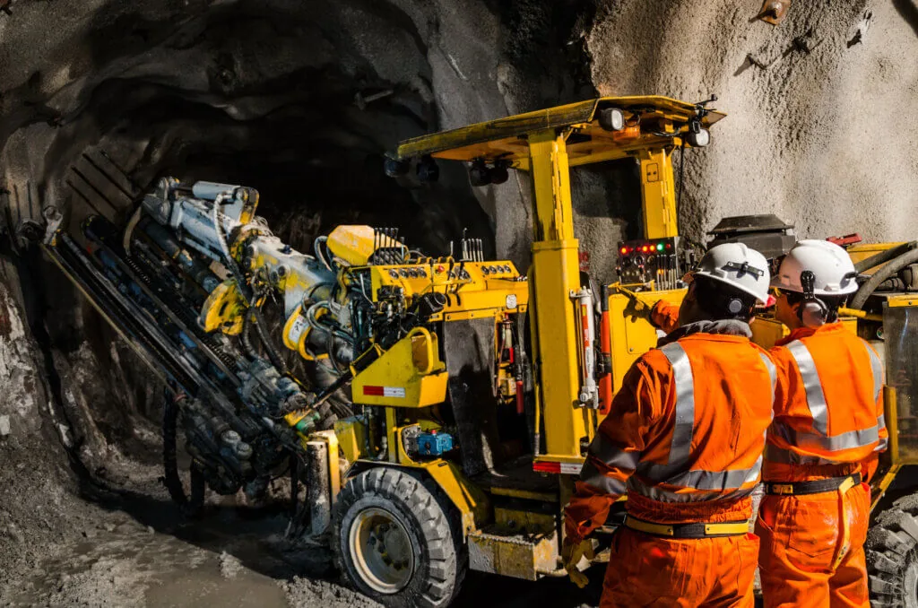 Two workers in orange safety suits and helmets operating a large drilling machine inside a mining tunnel.