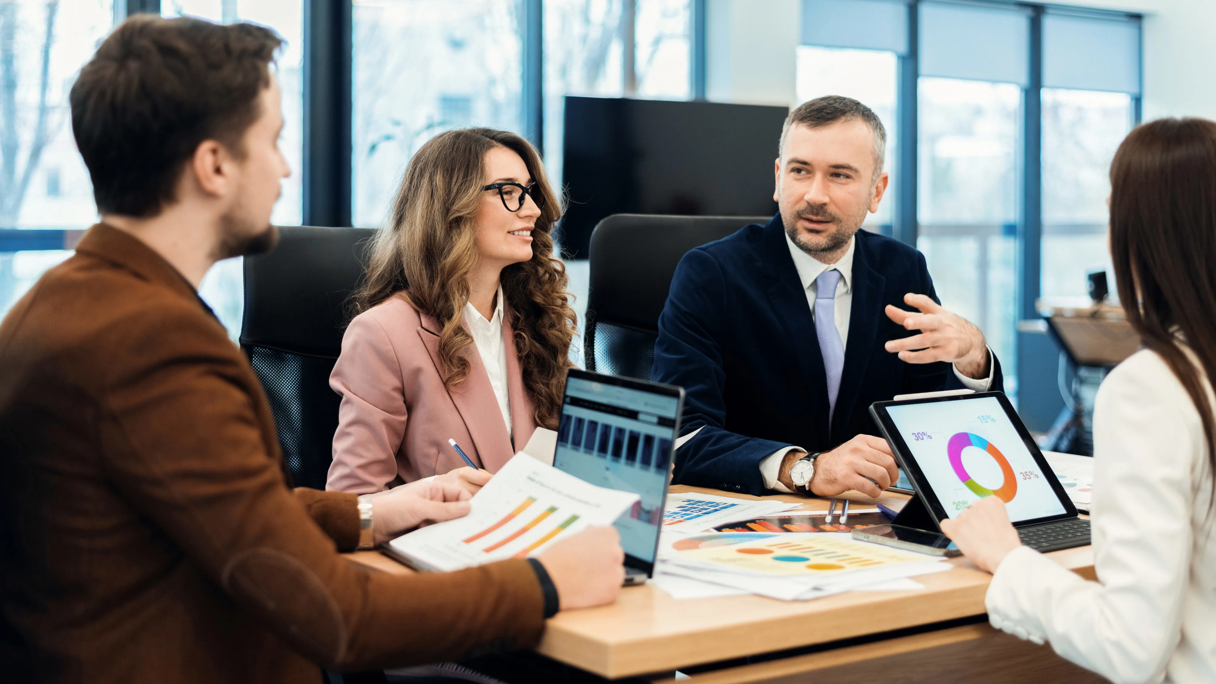 Four business professionals are seated around a conference table. A man with a beard in a suit is speaking and gesturing with his hand, while a woman with curly hair and glasses listens attentively. Laptops and papers with colorful charts are on the table.
