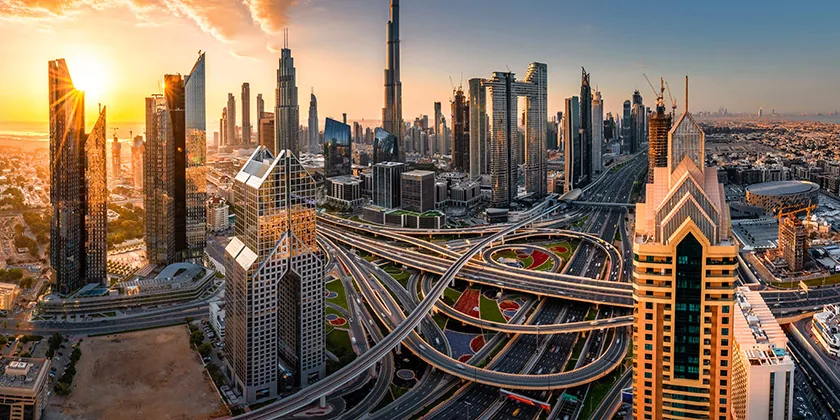 Panoramic cityscape of a major city with tall skyscrapers, intricate highways, and the sun setting in the background.