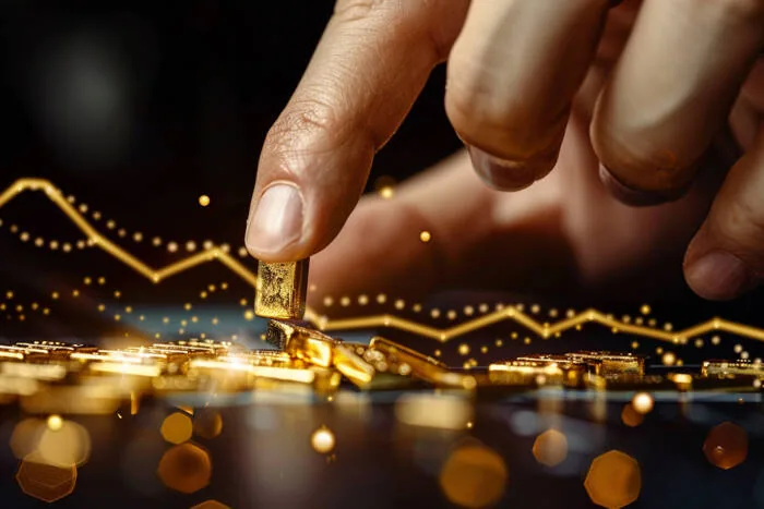Hand placing a small gold bar on a pile of gold with a financial chart overlay.