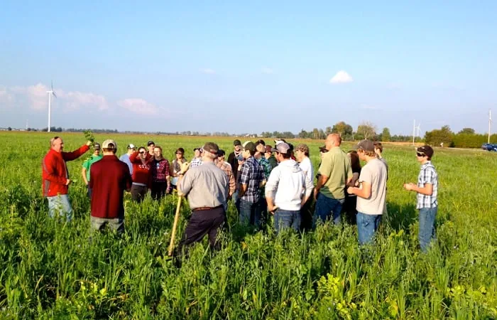 A group of people, including farmers and what appear to be agricultural students, are gathered in a green field listening to an instructor in a red jacket who is pointing at something in the field. Wind turbines are visible in the distance.