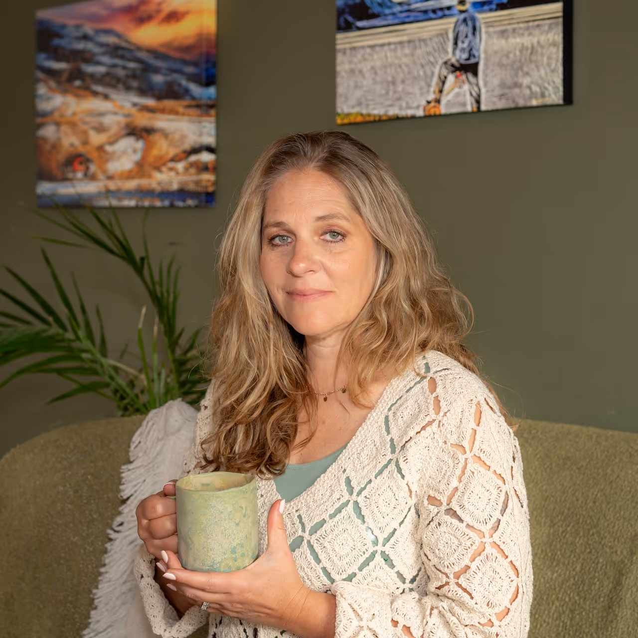 Woman with long wavy blonde hair sitting on a green couch, holding a green ceramic mug and wearing a cream crocheted cardigan.