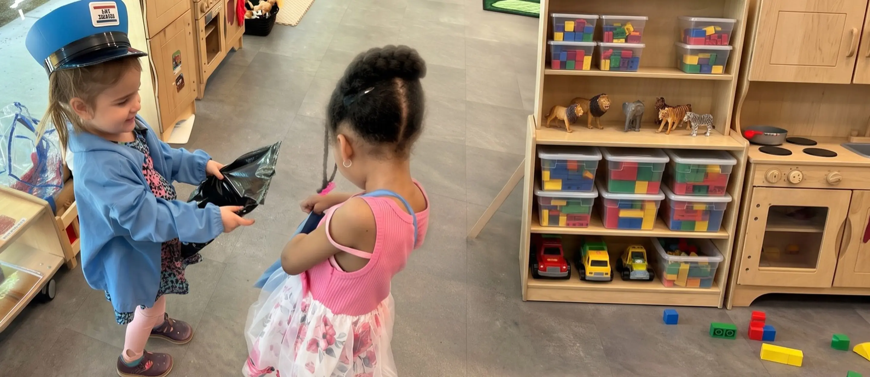 Two pre-K children playing dress-up in their classroom, one wearing a mail carrier hat