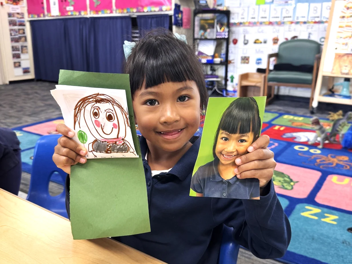 A smiling pre-K child holding up a self-portrait drawing next to their own photo