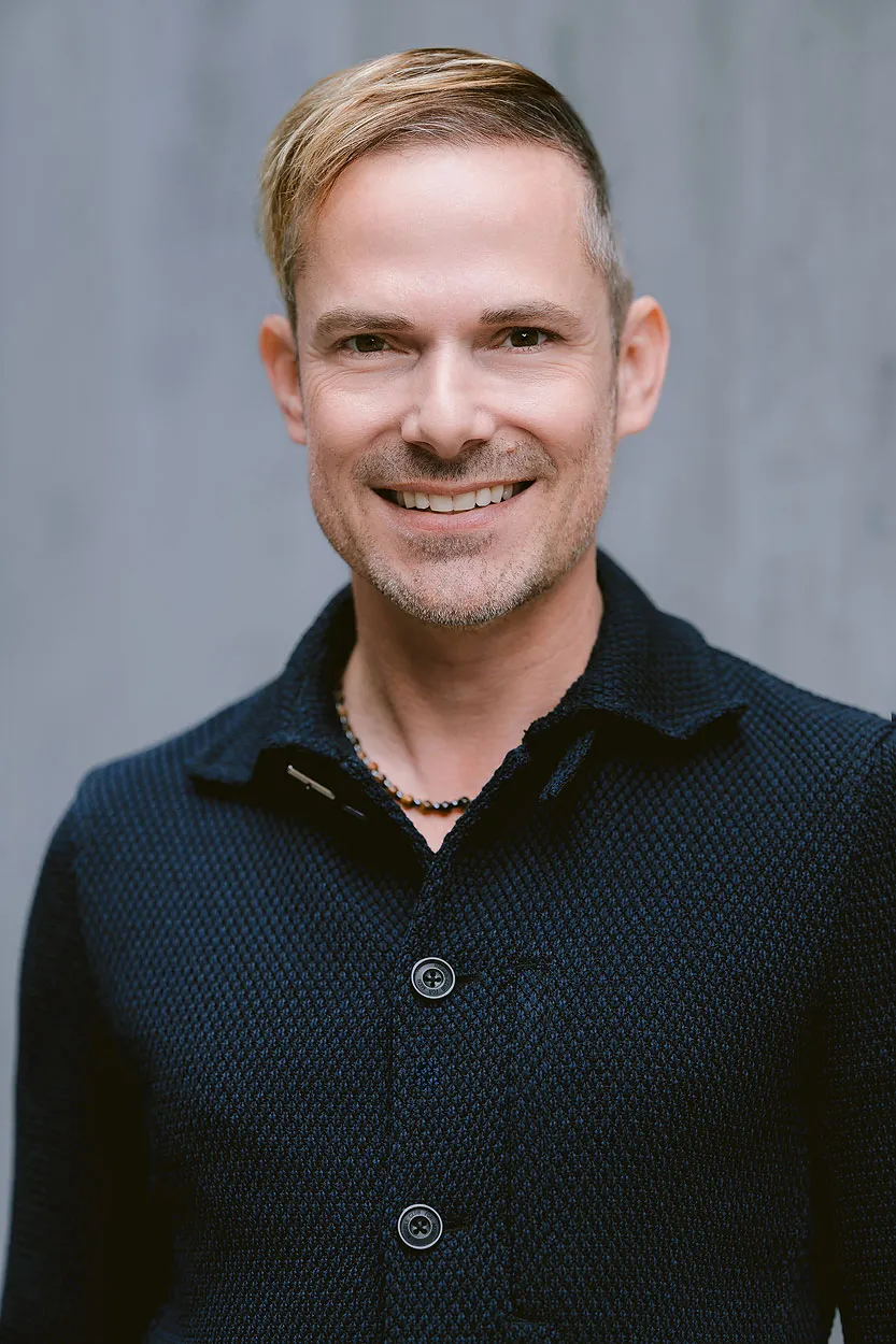 Adam Welch, founder of The Head Concierge, smiling in a dark textured jacket against a grey background