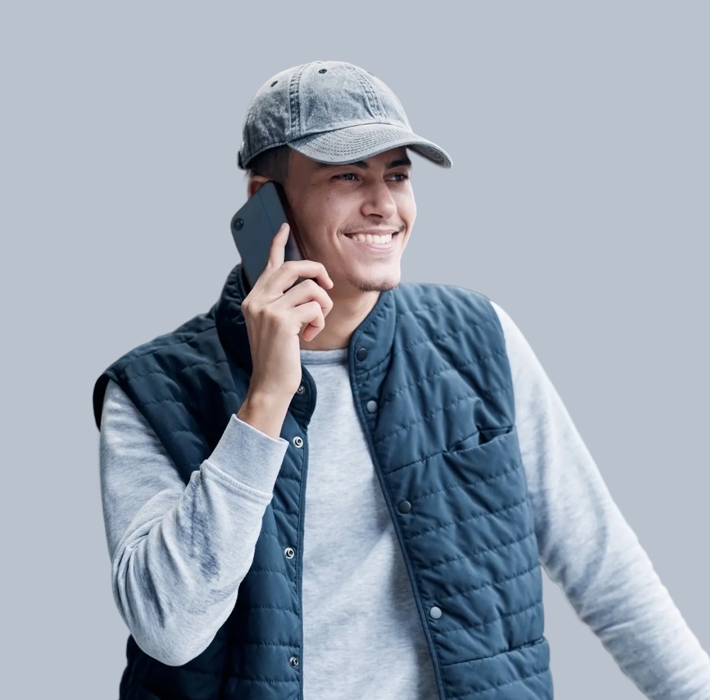 Smiling young man wearing a gray cap and navy blue quilted vest talking on a smartphone against a gray background.