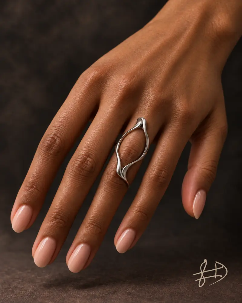 Main à la peau brune, aux ongles nude en amande, portant une bague longue argentée et ajourée sur le majeur ; signature claire en bas à droite.