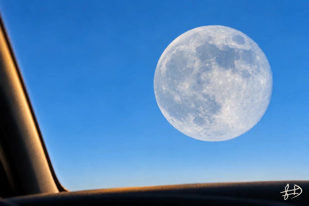 PROXIMITÉ : une immense pleine lune blanche et grise à droite d’un ciel bleu, vue depuis une voiture éclairée par le soleil de fin d’après-midi. Signature originale FAII en bas à droite.