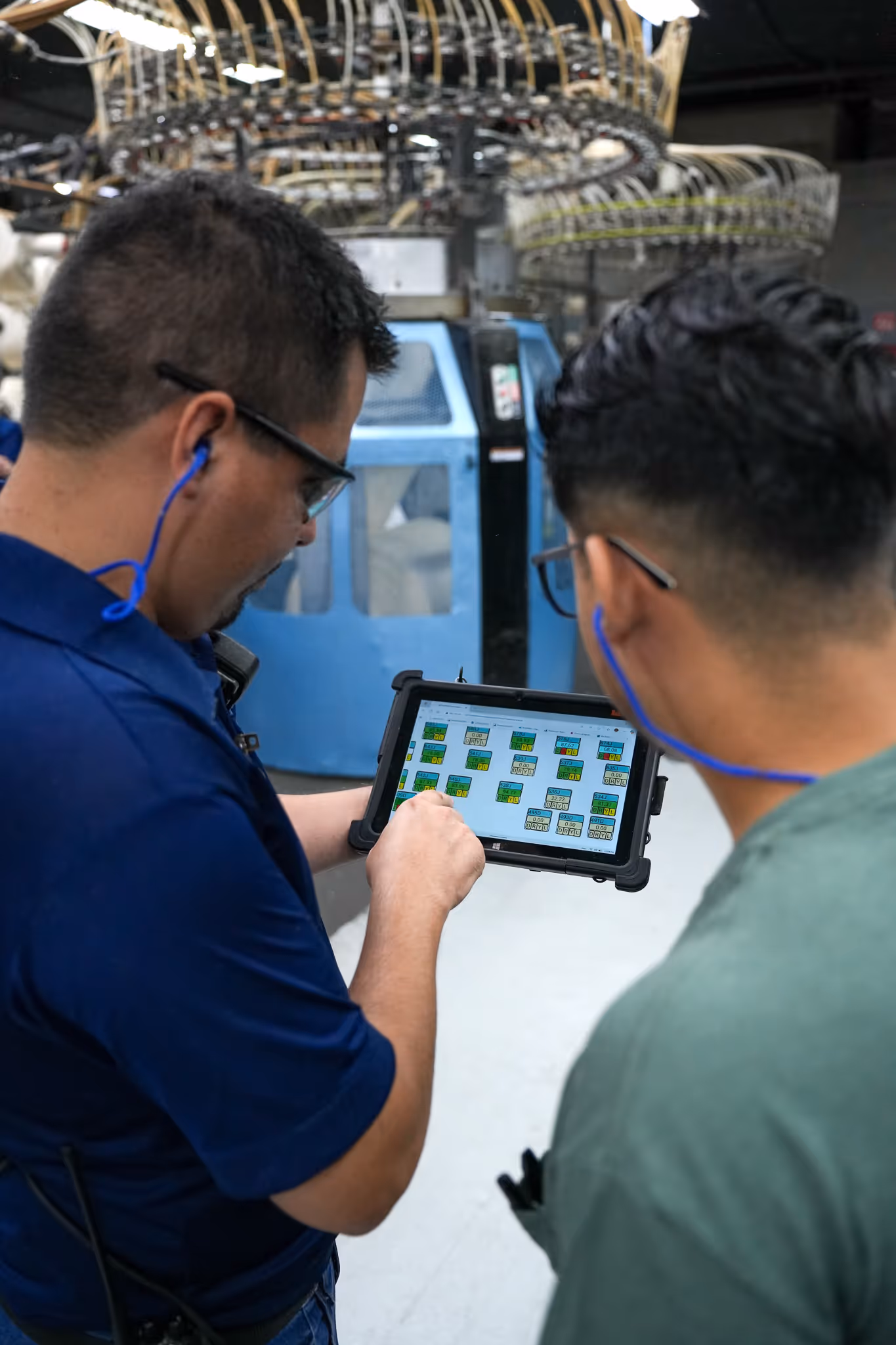 Two factory workers wearing safety glasses and earplugs viewing a digital control panel on a tablet inside an industrial setting.