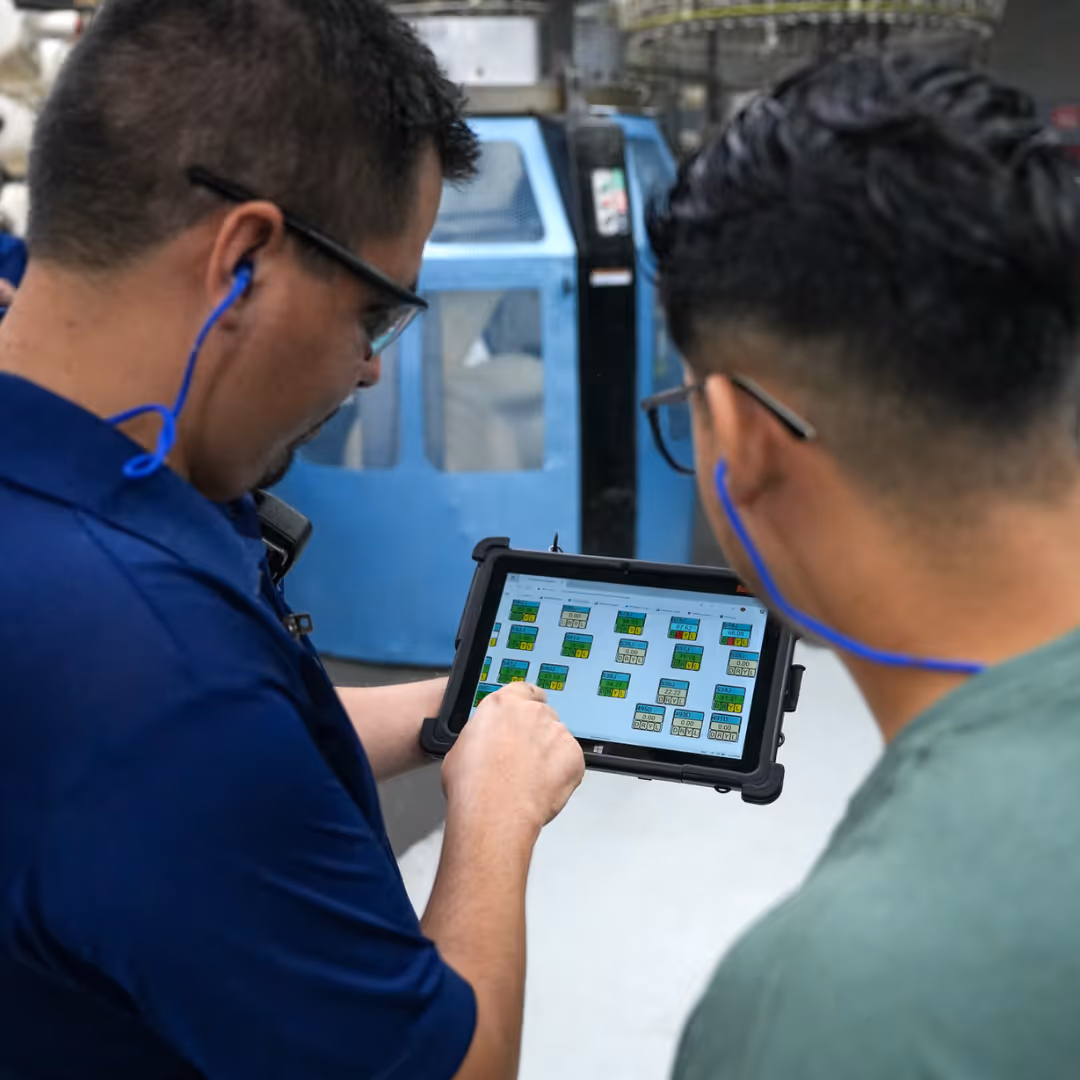 Two men wearing glasses and blue earphones looking at a tablet displaying a control panel interface in an industrial setting.