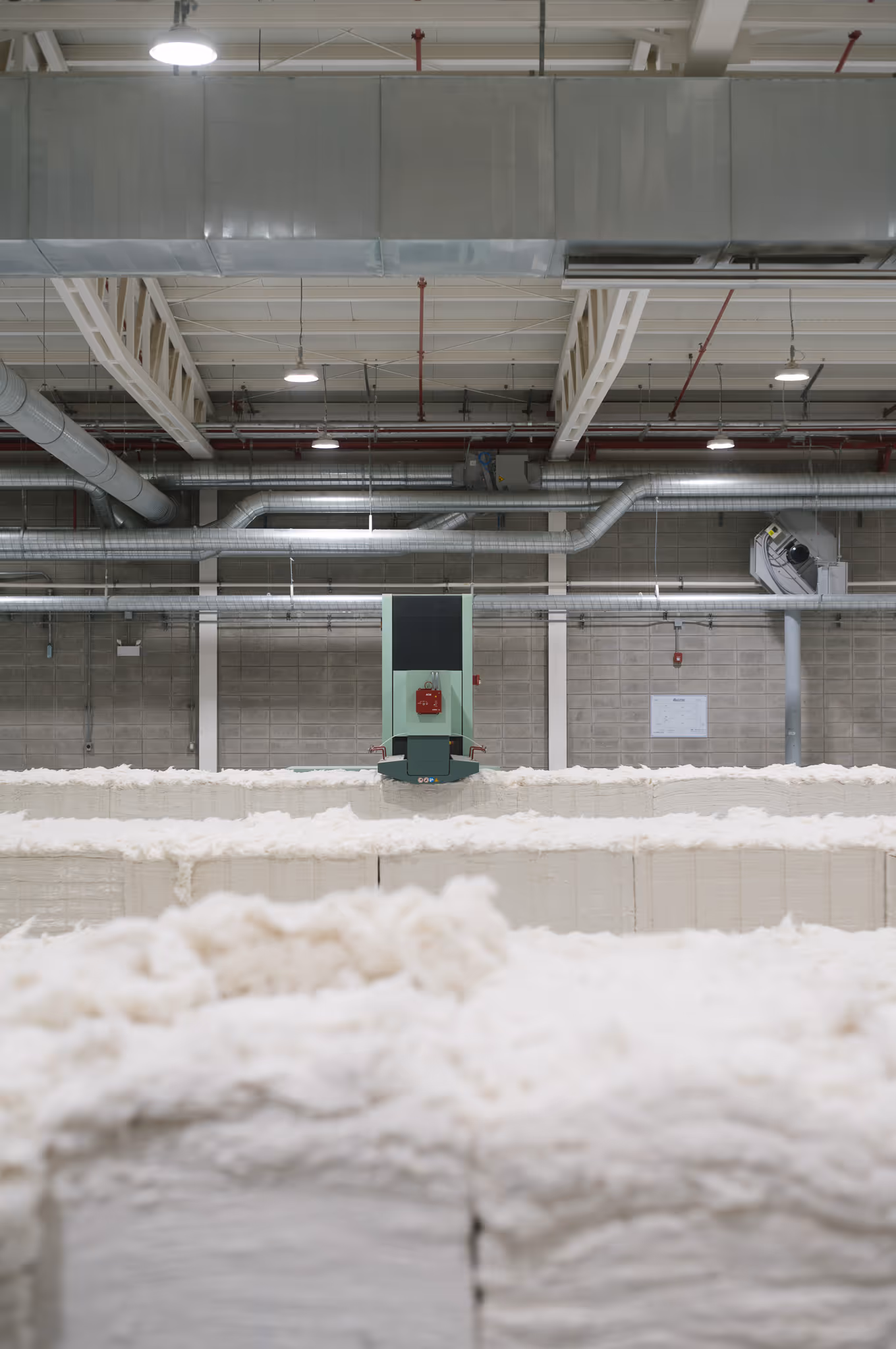 Industrial cotton processing facility with large stacks of raw cotton bales and overhead ventilation pipes.