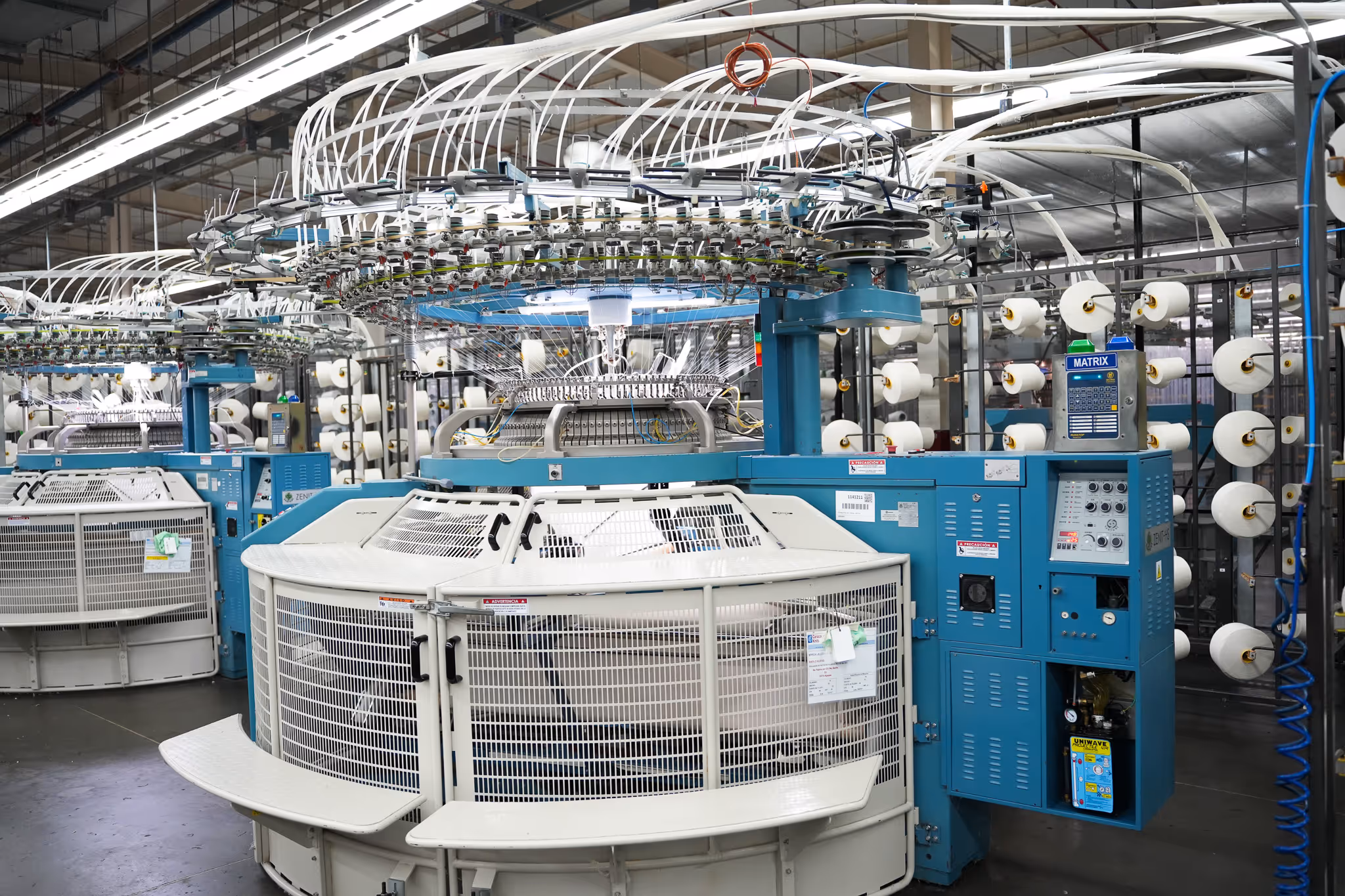 Industrial circular knitting machines with blue and white components in a textile factory.