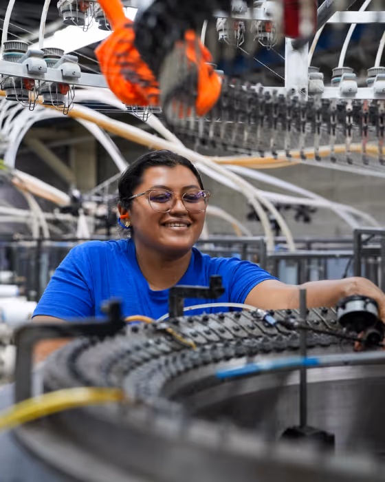 Smiling factory worker wearing glasses and a blue shirt operating machinery with curved conveyor belts and pipes in an industrial setting.