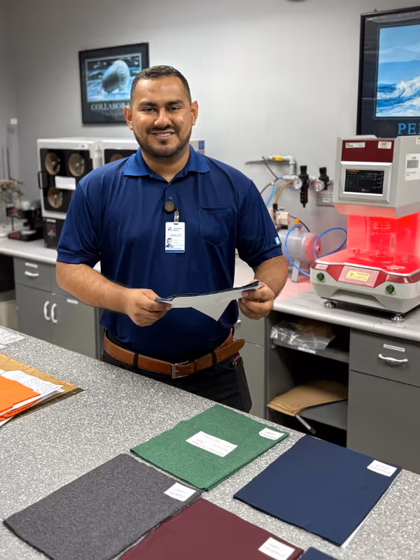 Man in blue polo shirt holding papers, standing behind a counter with various fabric samples in a laboratory or workshop setting.