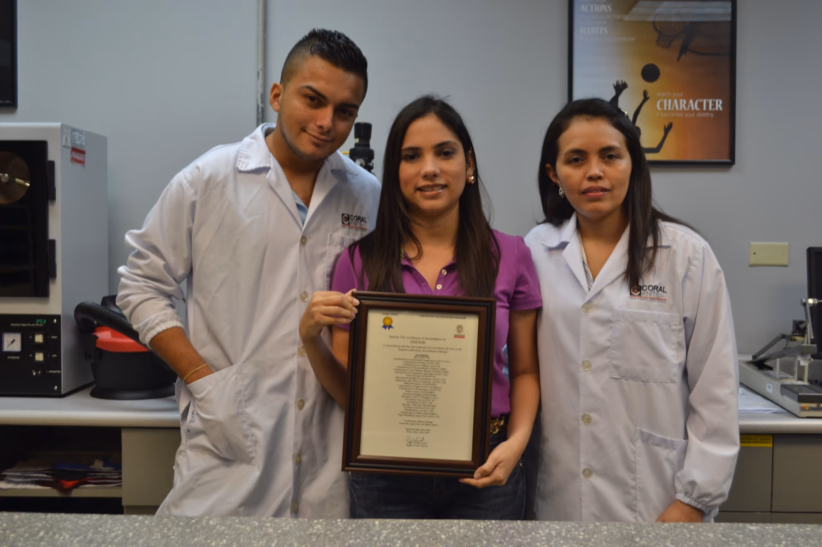 Three young adults, two wearing white lab coats and one holding a framed certificate, standing in a laboratory setting.