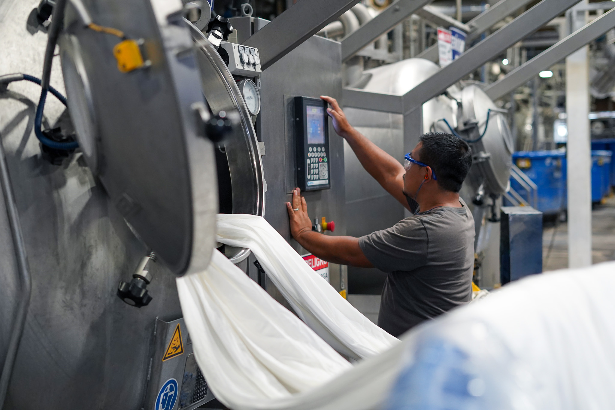 Factory worker operating industrial laundry machine with white fabric flowing out.