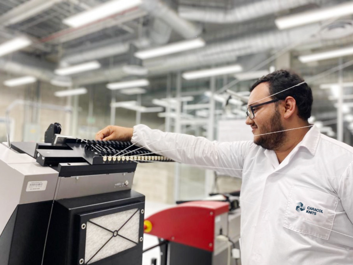 Technician wearing glasses and a white Caracol Knits lab coat adjusting a textile machine in a factory setting.
