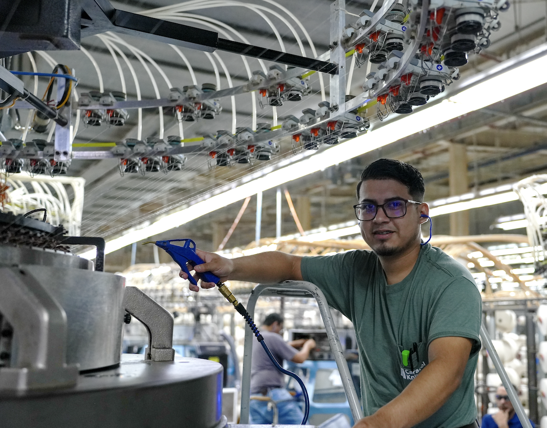 Factory worker wearing glasses and ear protection operating machinery with blue air blower in an industrial textile manufacturing setting.