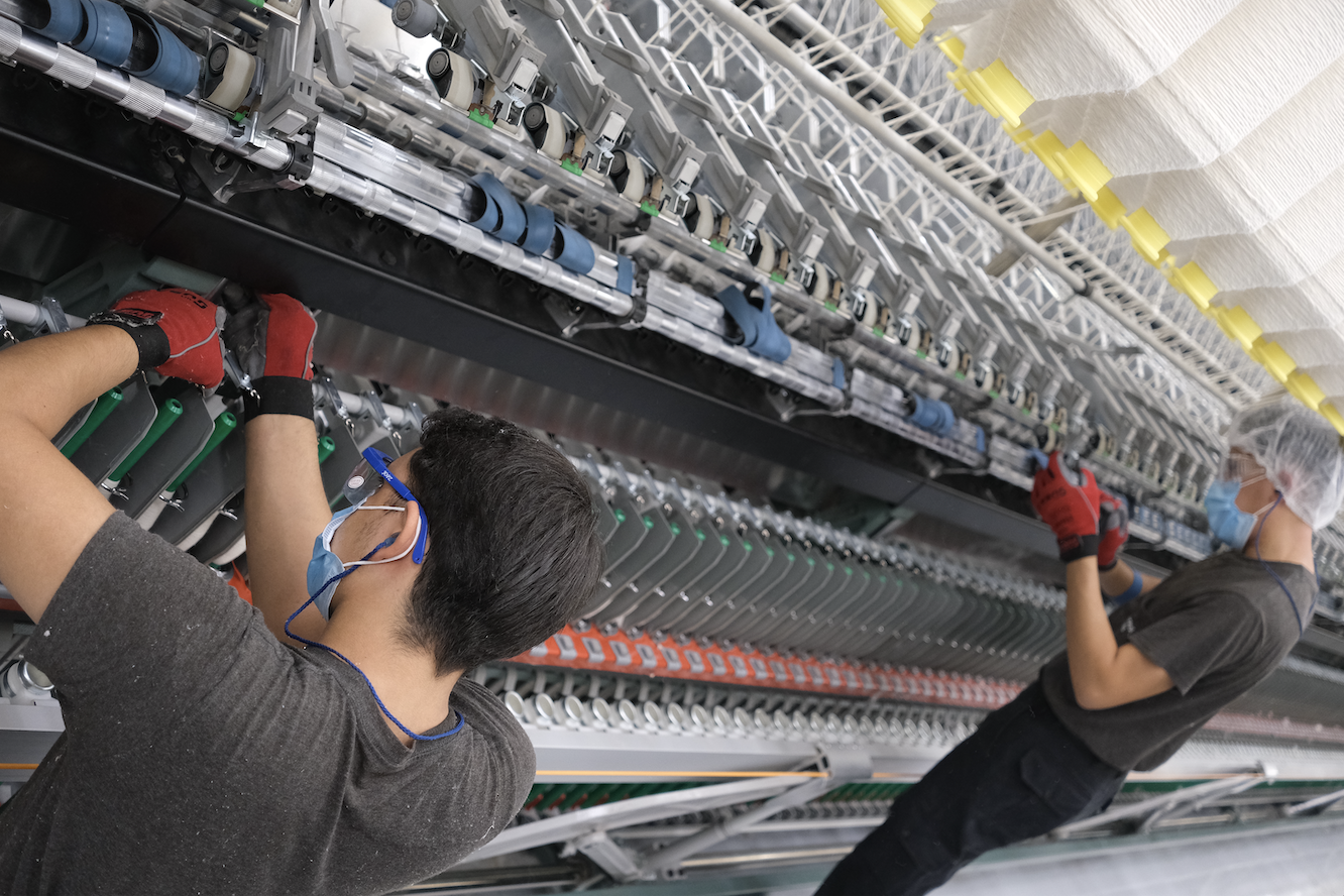 Two factory workers wearing masks and gloves operating a large industrial machine in a production facility.