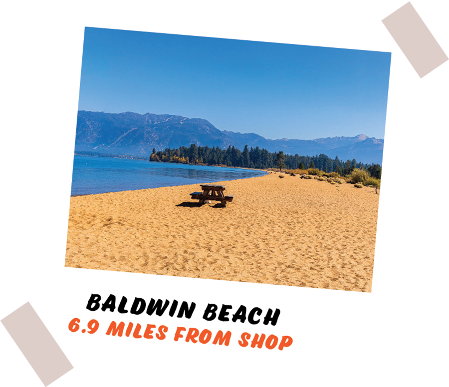 Sandy Baldwin Beach with a lone picnic table, calm blue water, pine trees, and distant mountains under a clear sky.