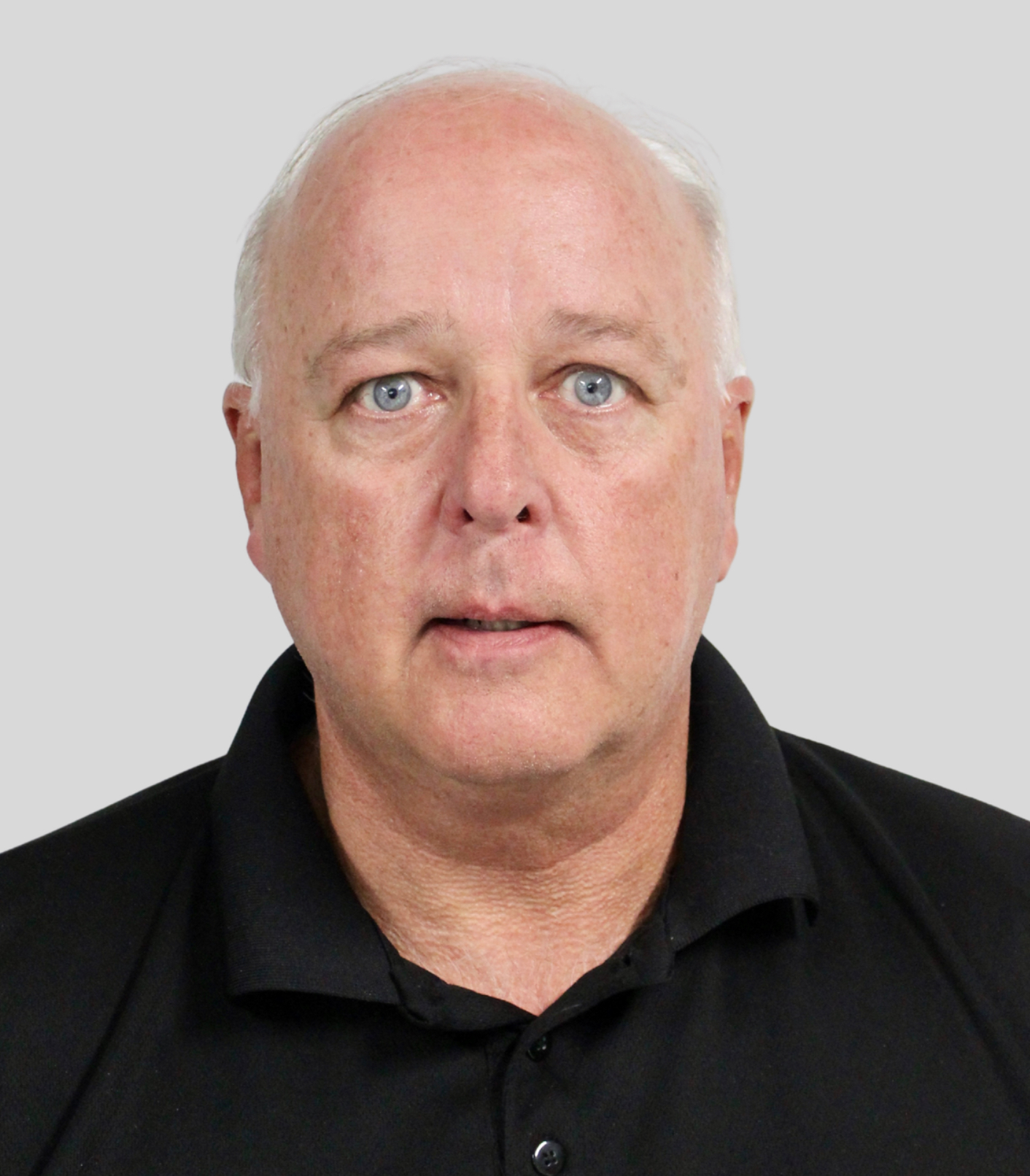 Portrait of Kevin with blue eyes and short gray hair wearing a black polo shirt against a plain gray background.