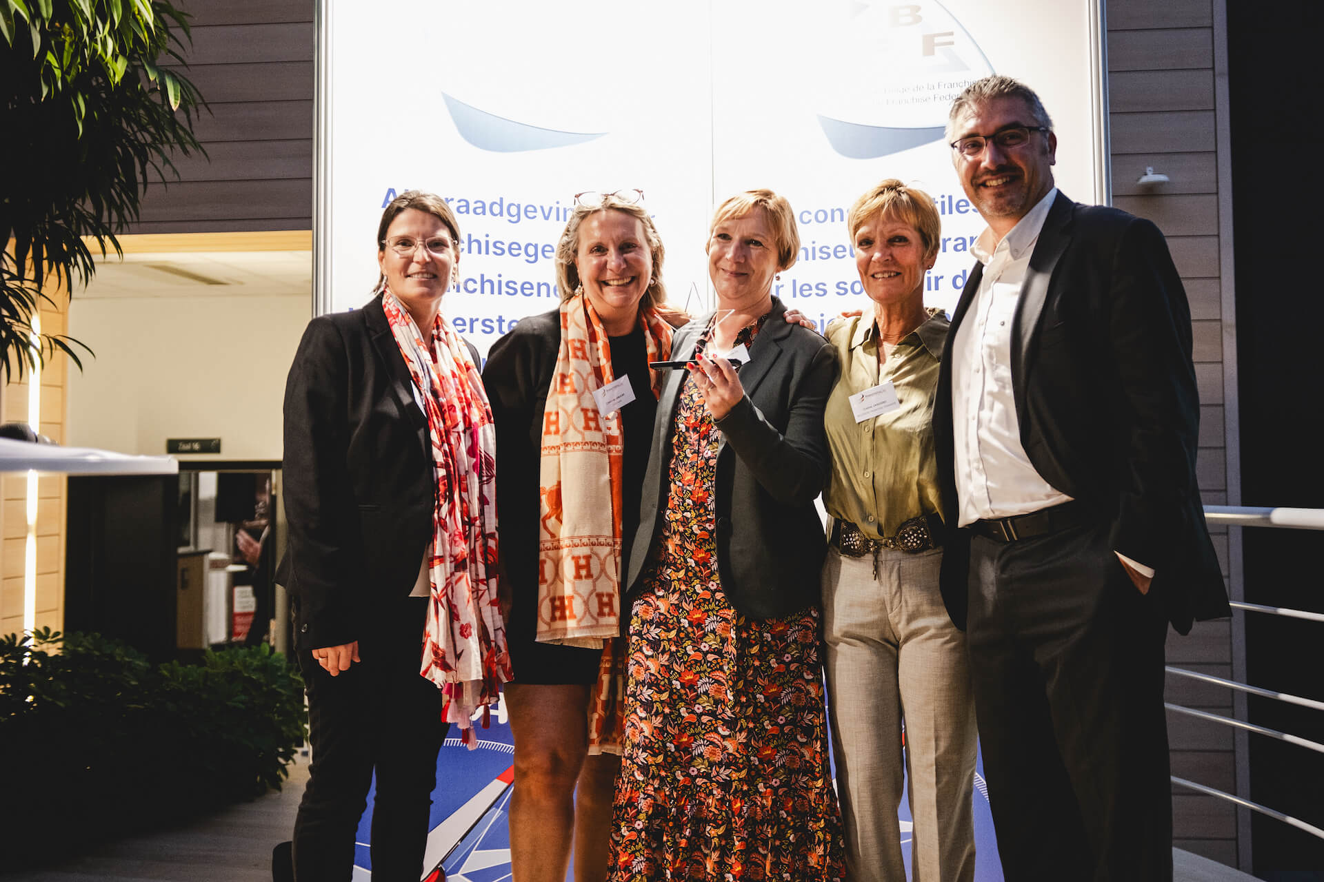 Cinq adultes souriants, quatre femmes et un homme en vêtements professionnels, posant ensemble devant un panneau informatif.