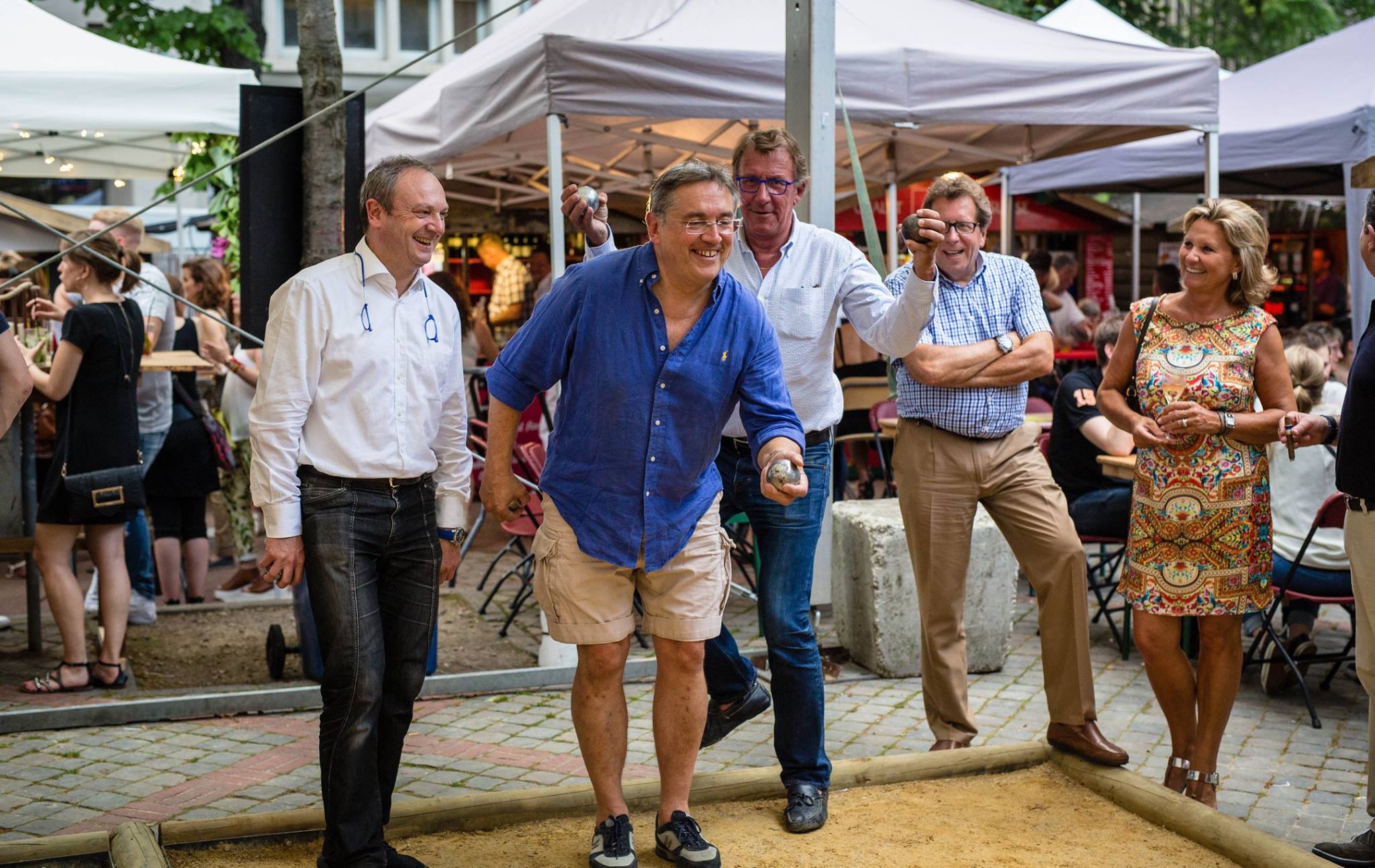 Groupe de cinq adultes jouant à la pétanque dans un cadre extérieur festif avec des tentes et des tables en arrière-plan.