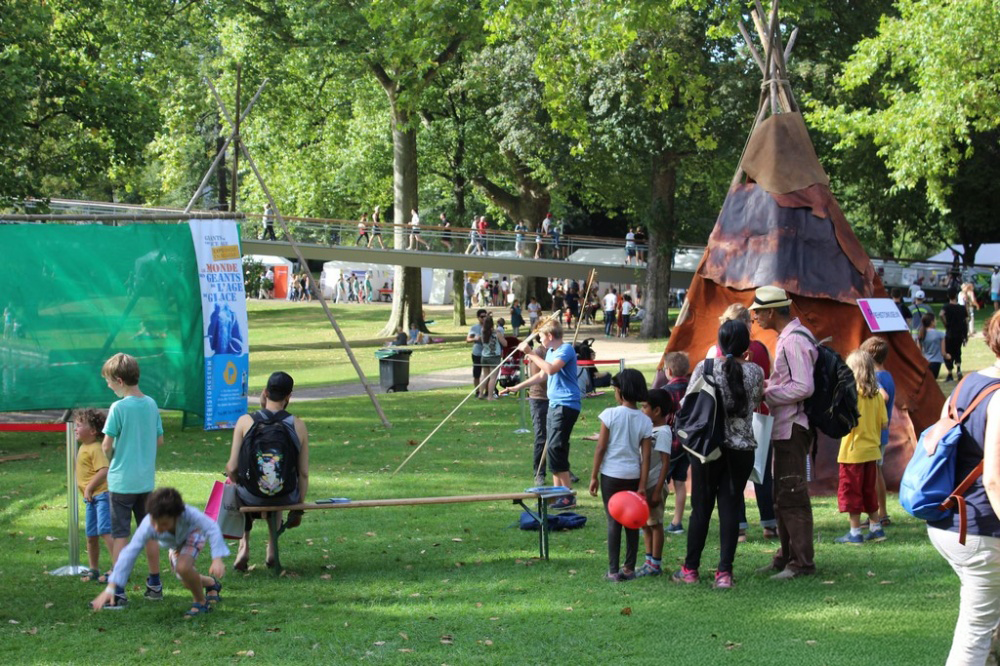 Groupe de personnes, principalement des enfants, participant à une activité en plein air dans un parc avec un tipi en toile et des arbres verts autour.