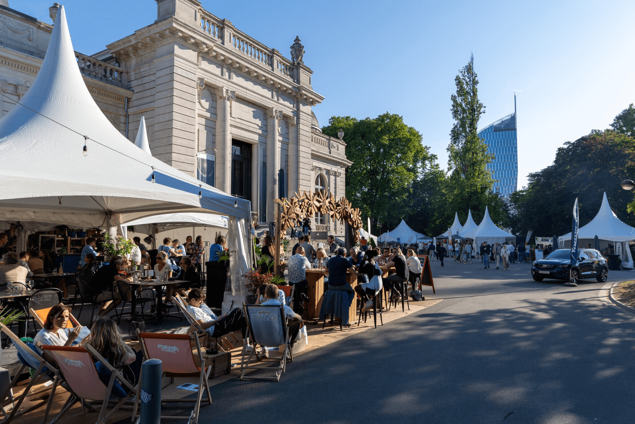 Personnes assises et socialisant sous des tentes blanches lors d'un événement en plein air près d'un bâtiment classique avec des arbres et un gratte-ciel en arrière-plan.