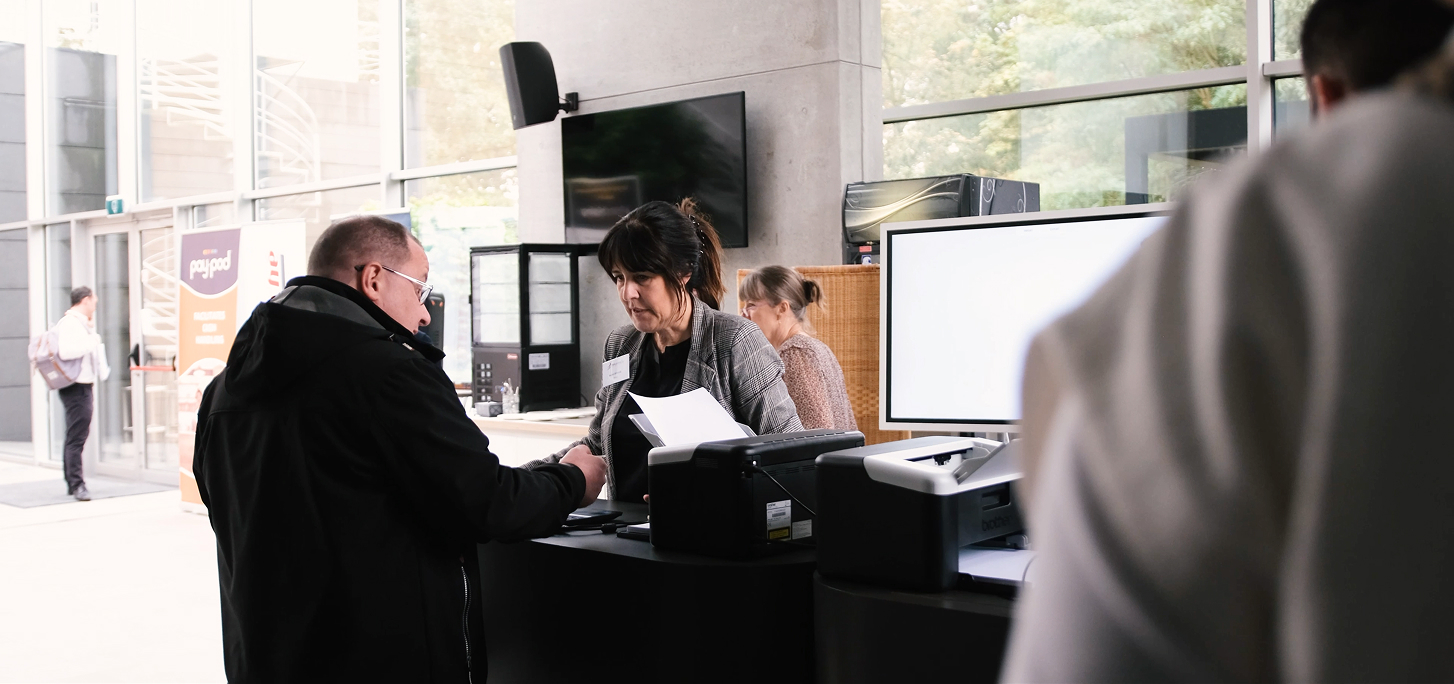 Deux femmes aidant des visiteurs à un comptoir d'accueil dans un bâtiment moderne avec des fenêtres larges.