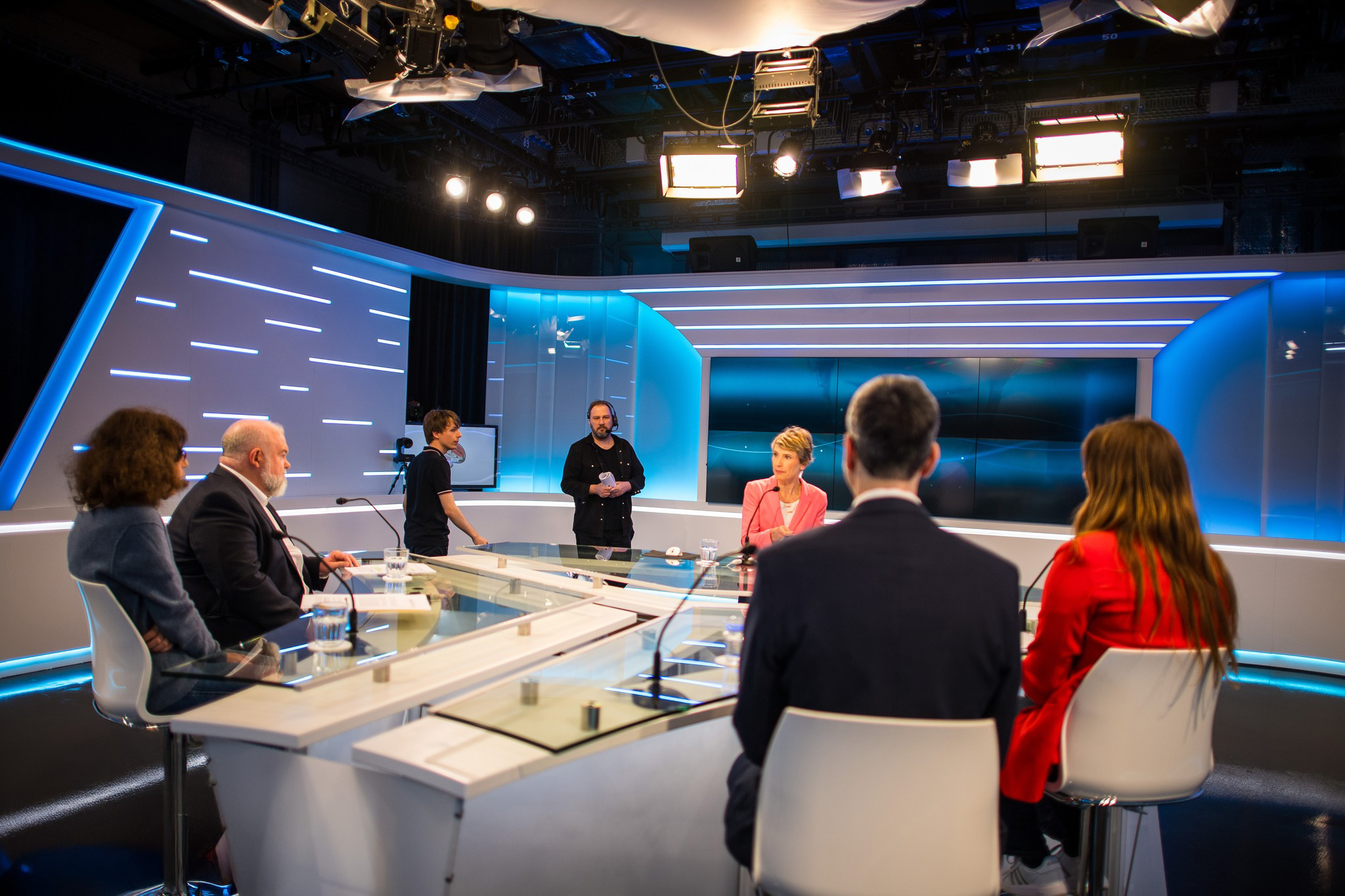Quatre personnes assises autour d'une table de studio télévisé moderne, deux techniciens debout en arrière-plan.