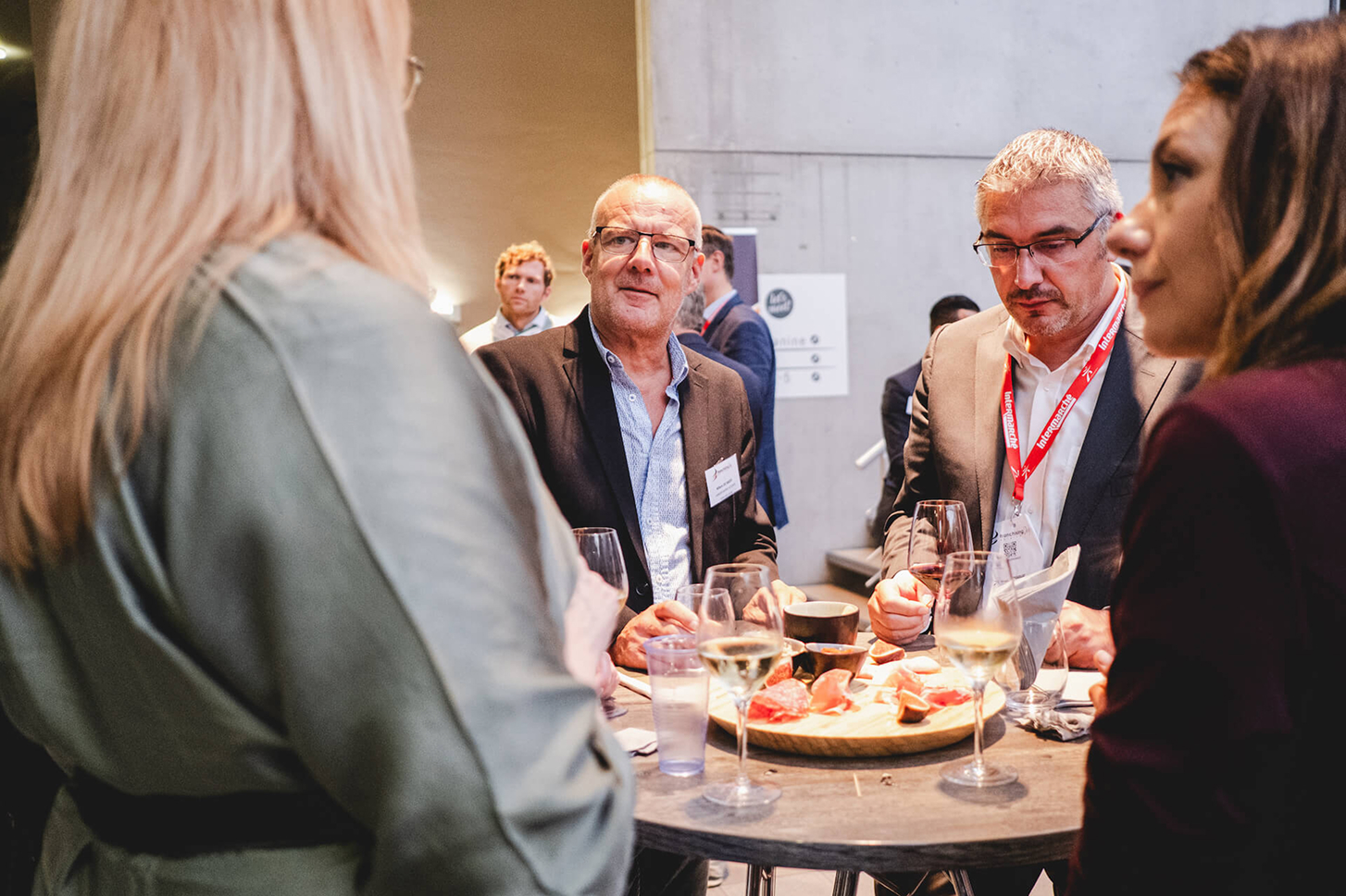 Quatre personnes en tenue professionnelle discutent autour d'une table haute avec charcuterie et verres de vin lors d'un événement.